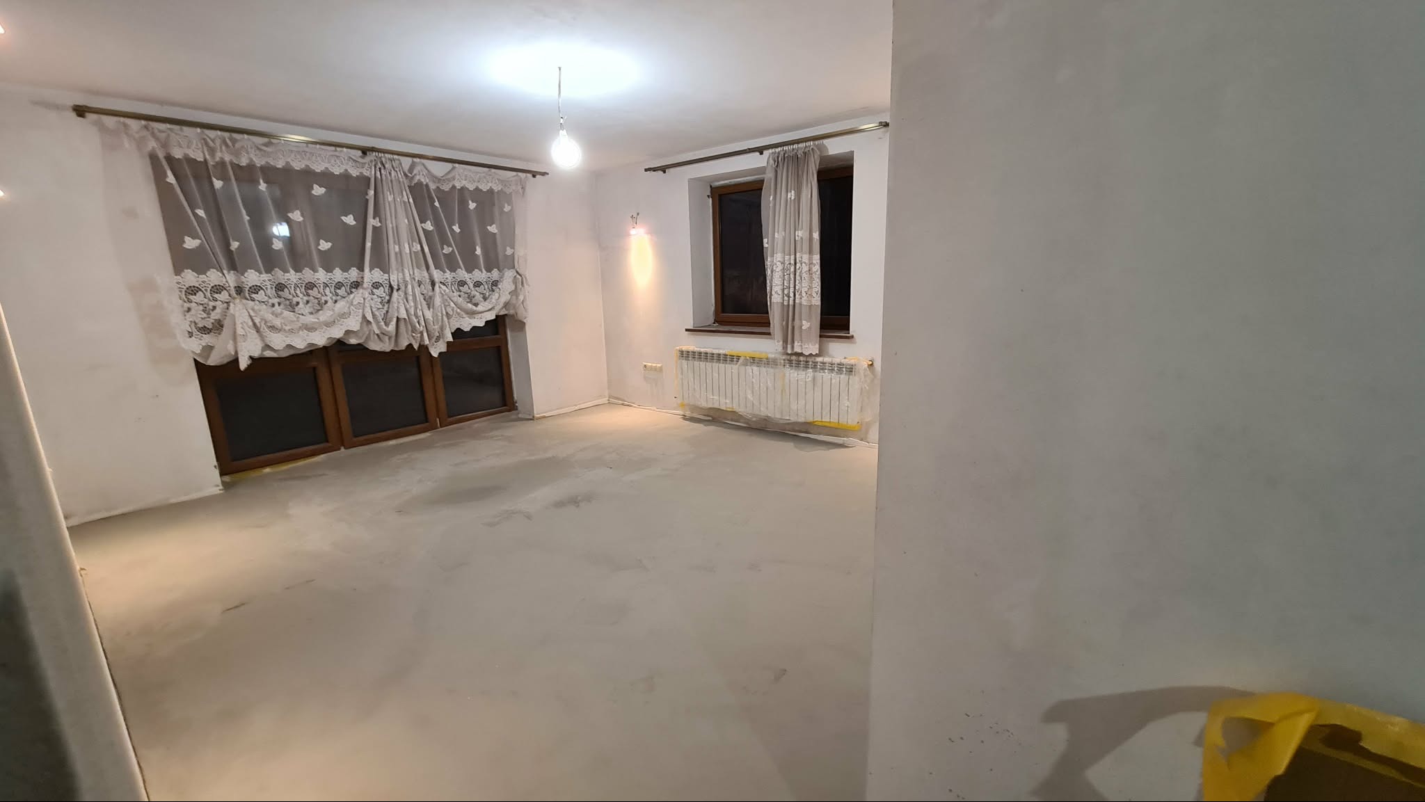 Pokój po wylewce, przygotowany do dalszych prac wykończeniowych. Okna z drewnianymi ramami, zasłony z koronkowym wzorem. Grzejnik zabezpieczony folią.