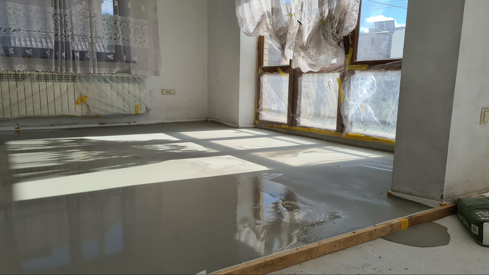 Wylany, świeży podkład podłogowy w jasnym pokoju z dużym oknem, zabezpieczonym folią. Widoczne drewniane listwy i zakryty grzejnik. Naturalne światło słoneczne.