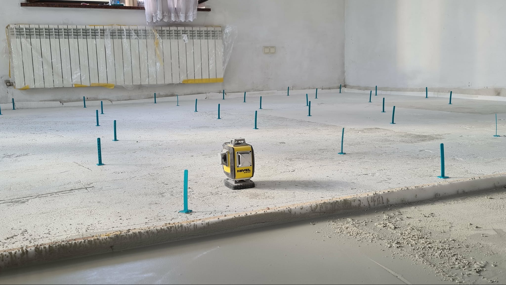 Wyrównywanie wylewki samopoziomującej z użyciem poziomicy laserowej i plastikowych bolców. Grzejnik zabezpieczony folią. Widok na szarą, mokrą powierzchnię.