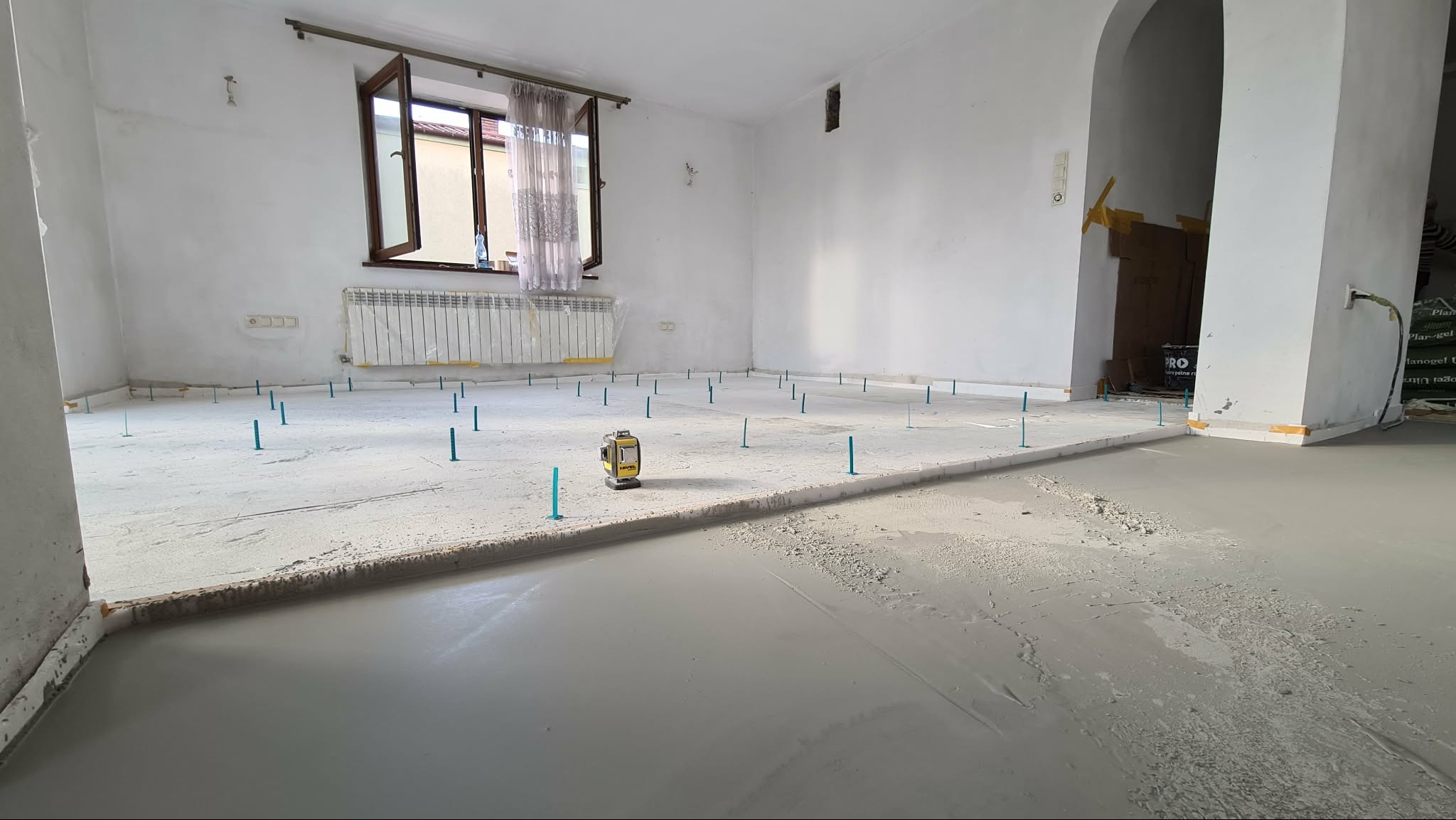 Wyrównywanie podłoża wylewką samopoziomującą z użyciem poziomicy laserowej i plastikowych bolców. Widok na okno z zasłonami i przejście w łuku.