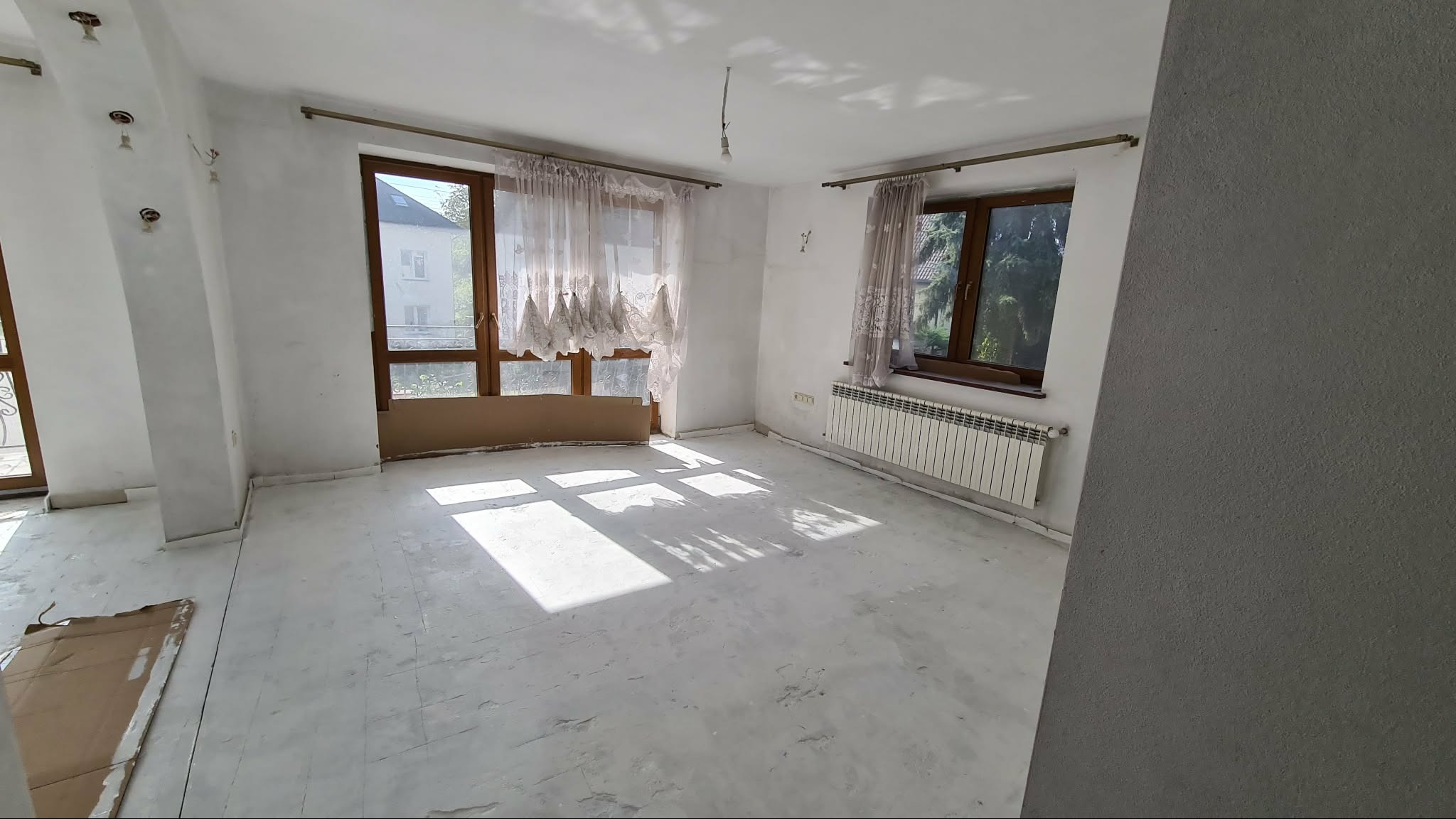Pokój przed remontem: surowe ściany, okna z firankami, grzejnik. Widoczne ślady po instalacjach i niedoskonałości podłogi. Przygotowanie do prac wykończeniowych.