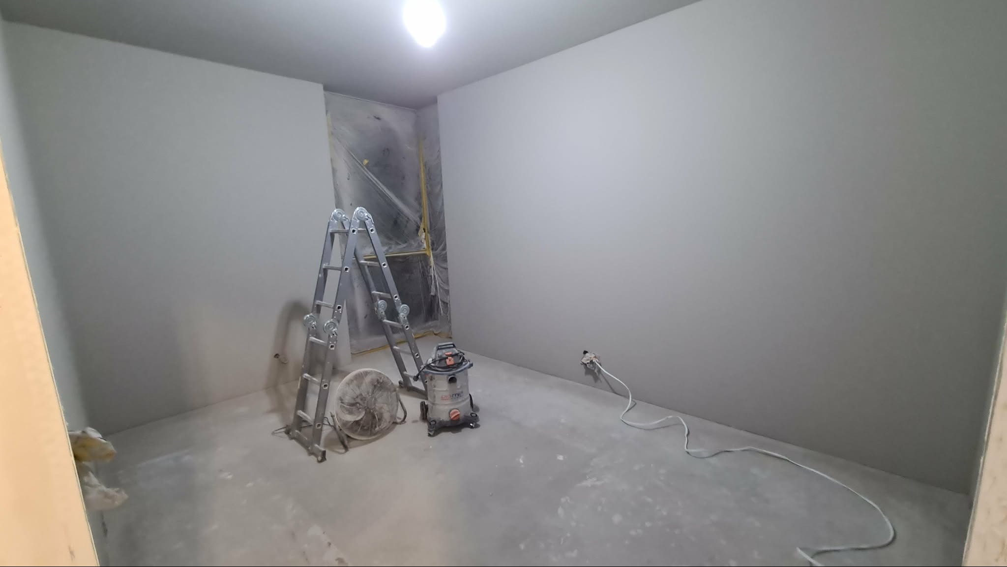 Wnętrze po szpachlowaniu: gładkie, szare ściany, drabina, wentylator i odkurzacz przemysłowy. Okno zabezpieczone folią. Widoczne gniazdko elektryczne.