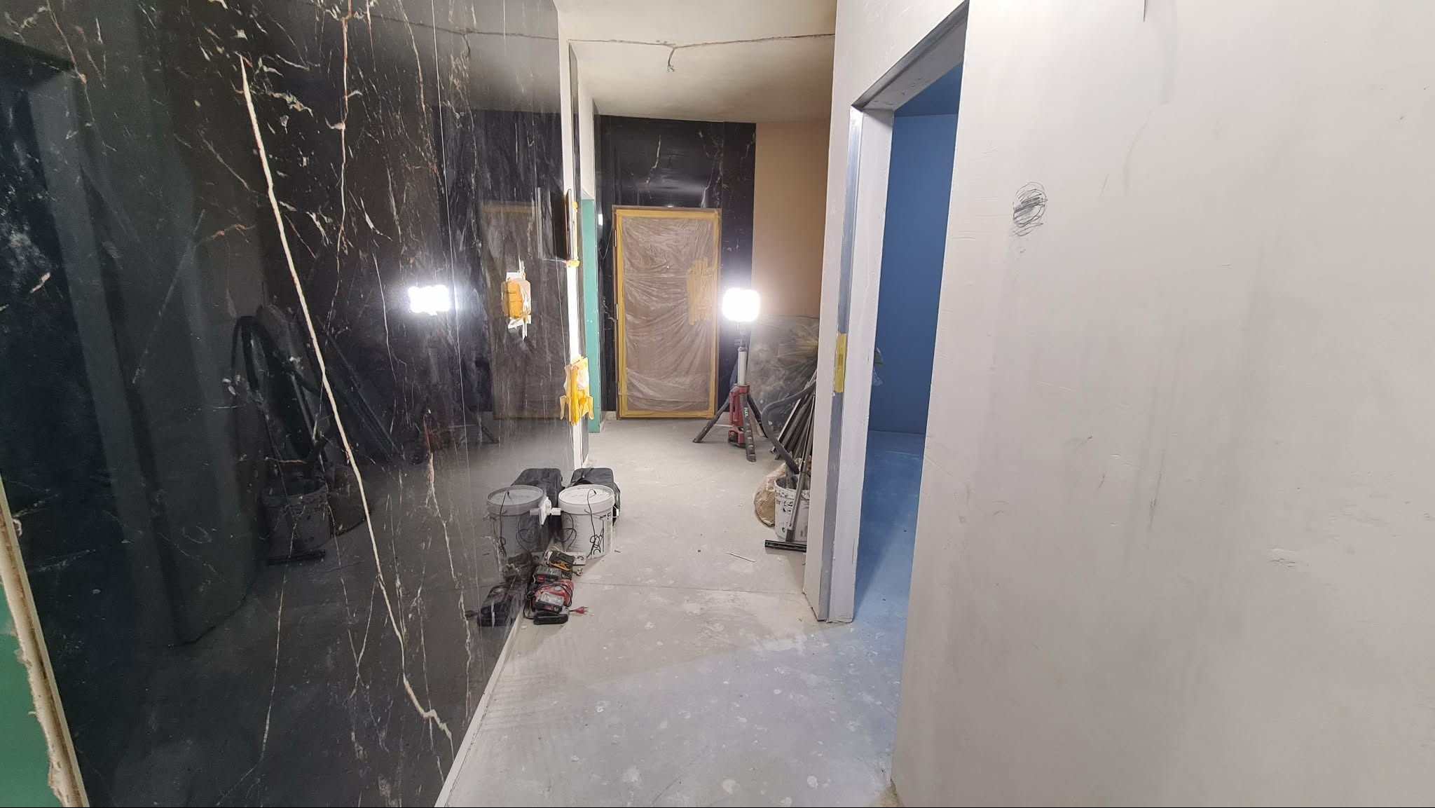 Wnętrze w trakcie wykańczania: czarne marmurowe płytki na ścianie, zabezpieczone drzwi, widoczne narzędzia i lampa budowlana. Ściany przygotowane do malowania.