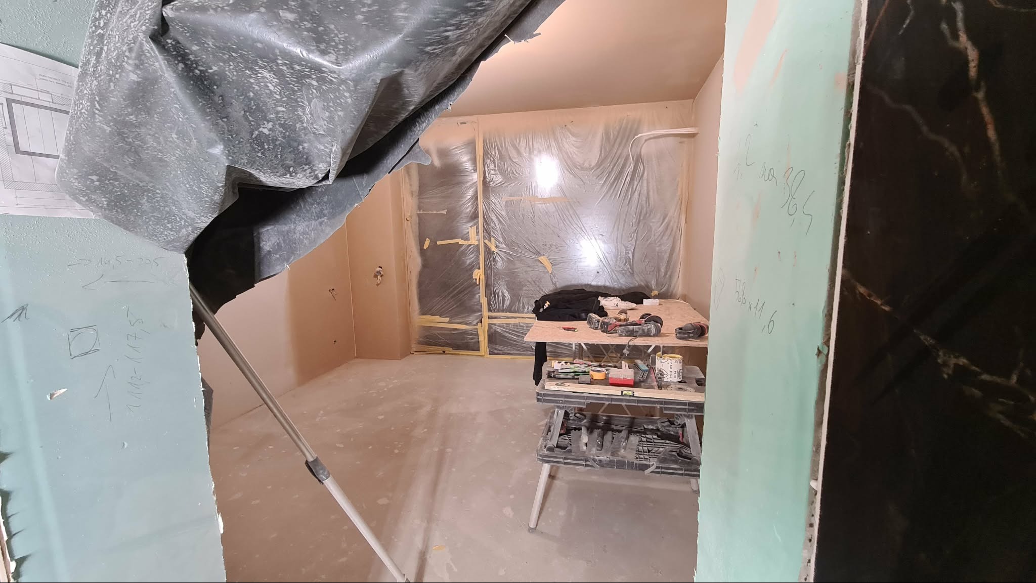 Pokój w trakcie prac wykończeniowych: zabezpieczone okna folią, narzędzia na stole warsztatowym, ściany z zapiskami, widoczne płyty karton-gips.