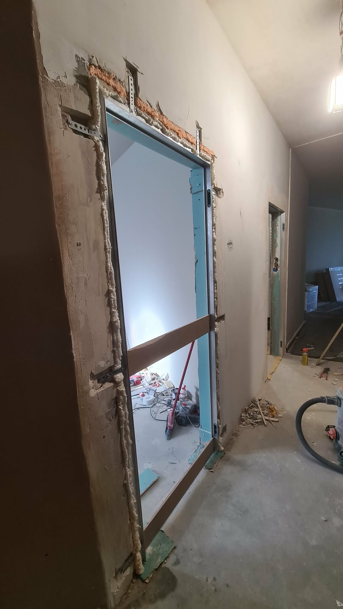 Montaż ościeżnicy metalowej z pianką montażową w surowym otworze drzwiowym. Widoczne narzędzia i resztki materiałów budowlanych. W tle kolejne pomieszczenie w trakcie prac.