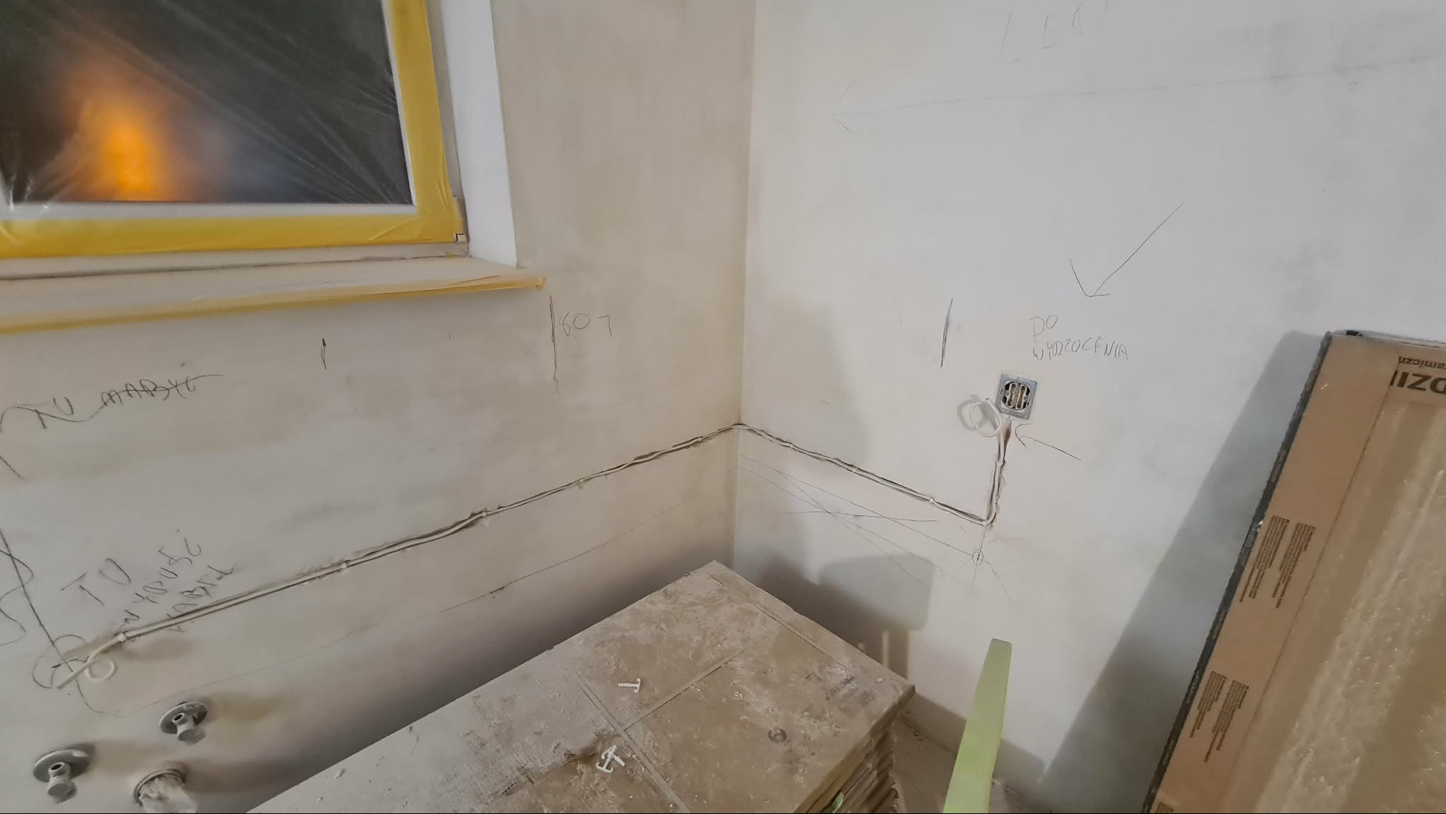 Ściana po szpachlowaniu z widocznymi przewodami elektrycznymi, przygotowana do dalszych prac wykończeniowych. Okno zabezpieczone folią malarską.