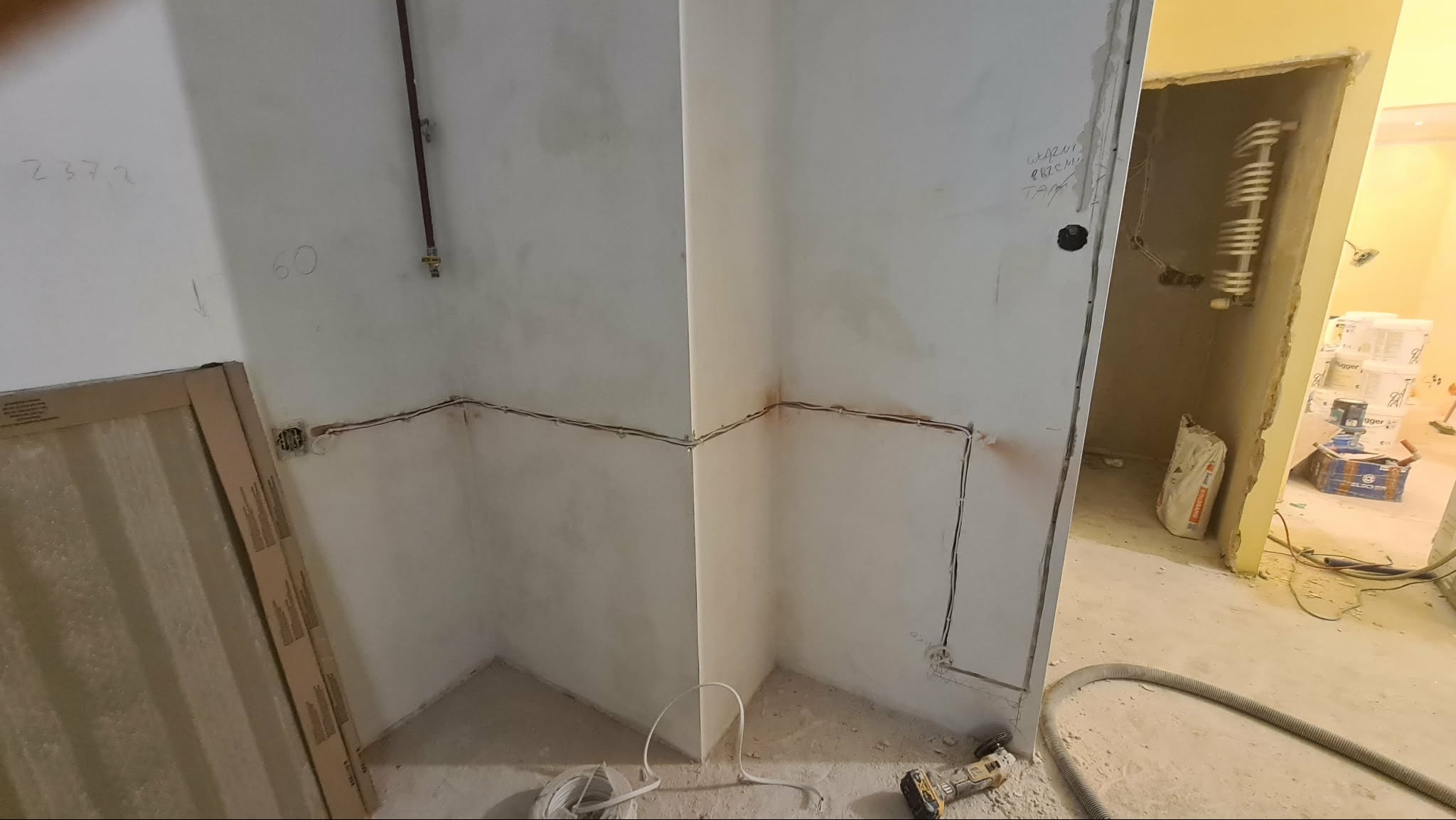 Wnętrze w trakcie prac wykończeniowych: widoczne kable elektryczne poprowadzone wzdłuż ścian, przygotowane do montażu gniazdek. W tle worki z zaprawą i narzędzia.