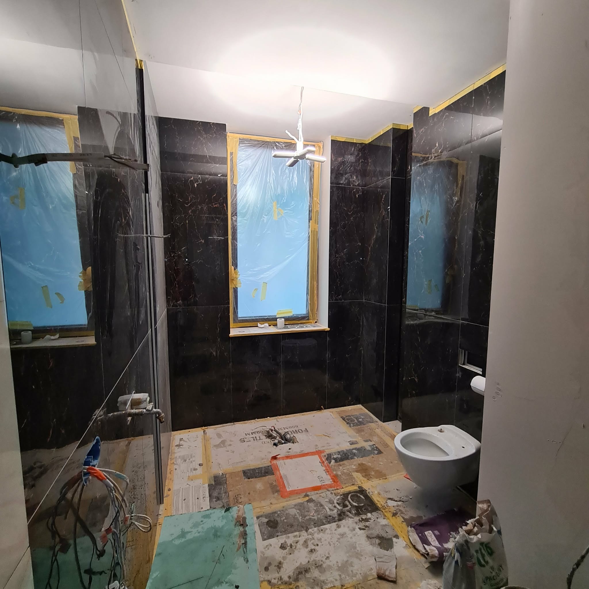Wnętrze łazienki w trakcie remontu. Czarne płytki imitujące marmur na ścianach, okno zabezpieczone folią, widoczne przygotowania do położenia podłogi i montażu toalety.