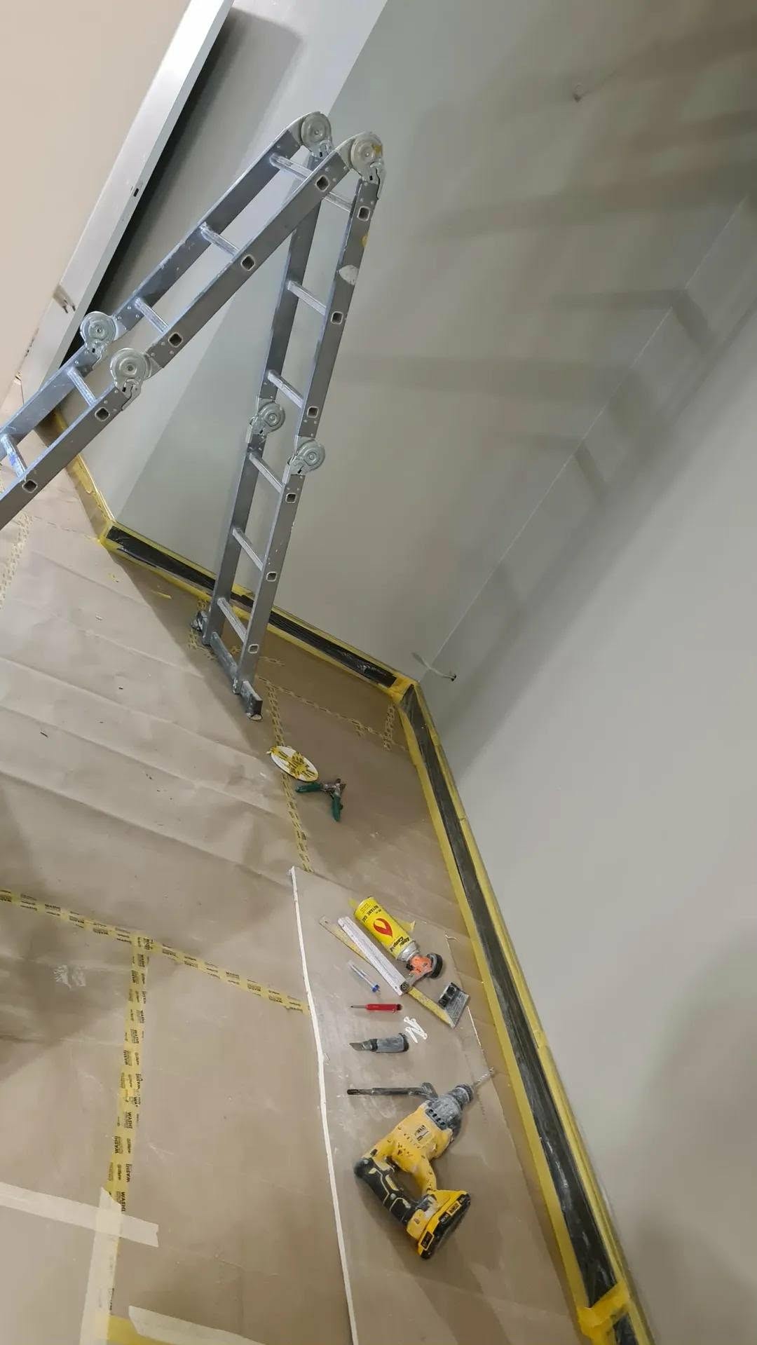 Świeżo pomalowane jasnoszare ściany klatki schodowej zabezpieczone taśmą malarską, z rozstawioną drabiną i narzędziami malarskimi na podłodze.