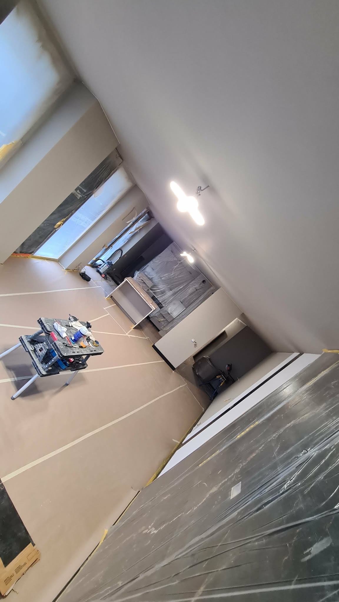 Wnętrze w trakcie prac wykończeniowych: zabezpieczona folią podłoga, narzędzia na stole warsztatowym, świeżo pomalowane ściany i sufit w jasnych barwach. Widok z góry.