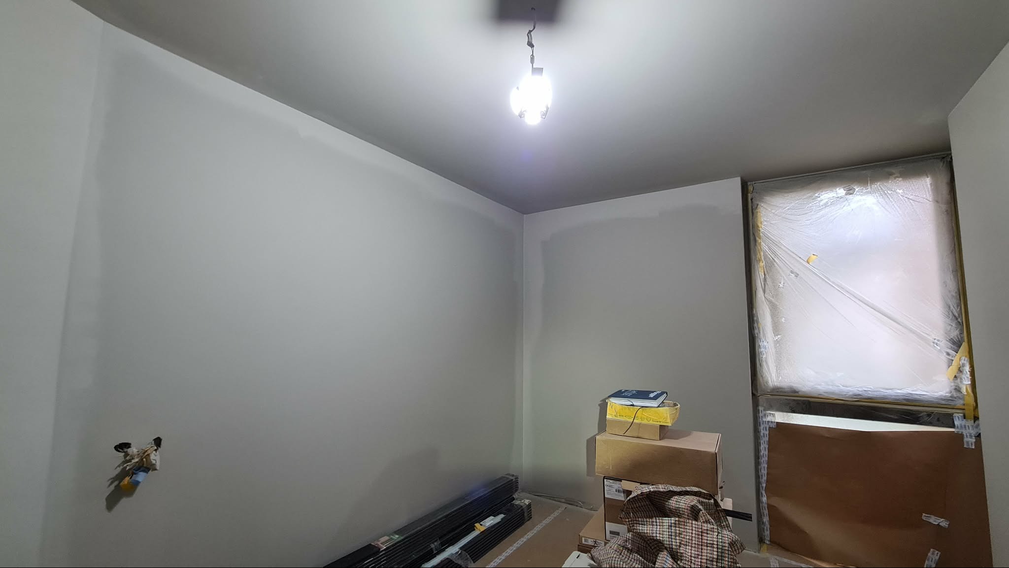 Pokój po szpachlowaniu ścian na gładko, przygotowany do malowania. Okno zabezpieczone folią malarską, widoczne materiały budowlane i wisząca żarówka.
