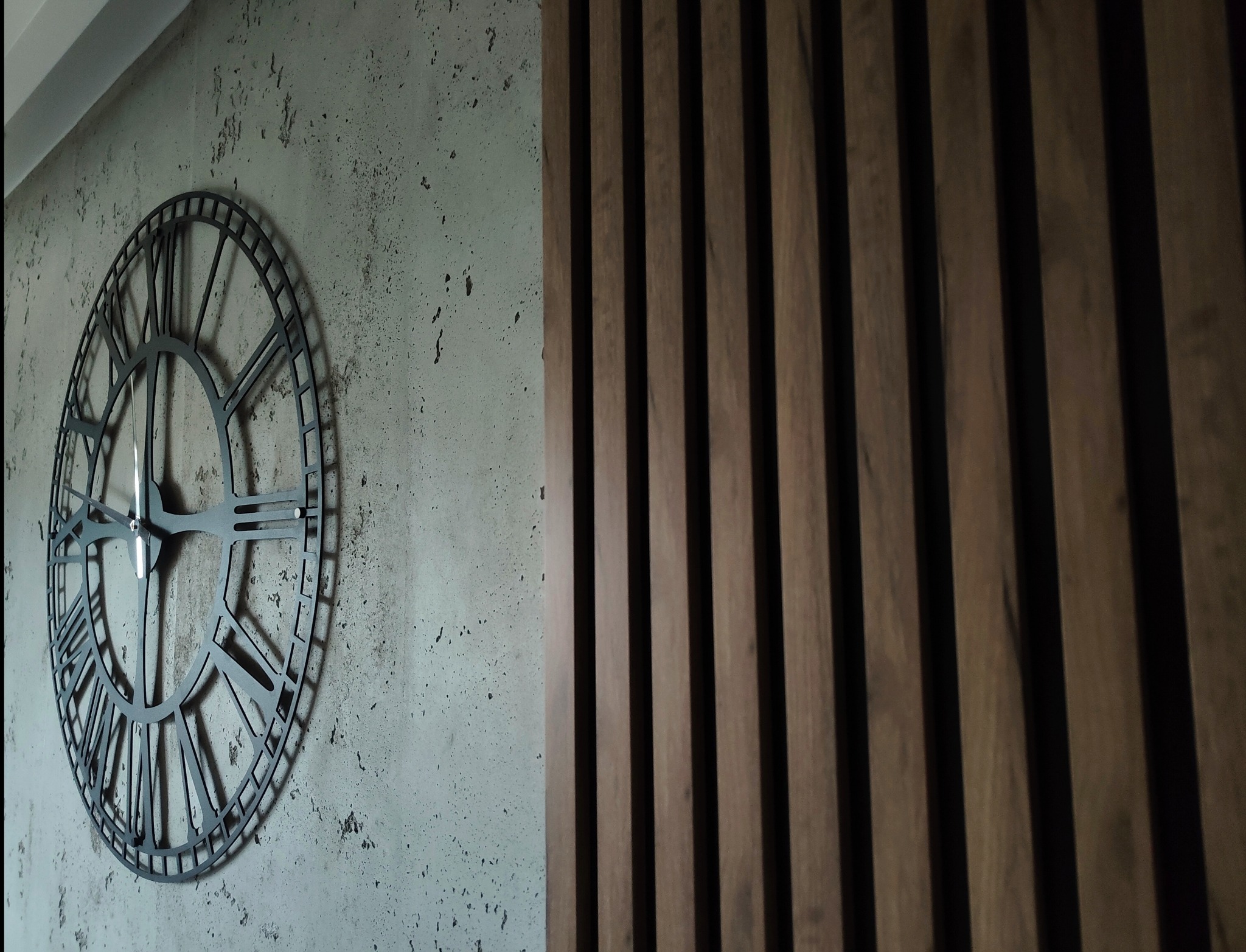 Dekoracyjna ściana z betonową fakturą i metalowym zegarem, obok pionowe drewniane lamele. Nowoczesny design wnętrza z elementami industrialnymi i drewnianymi.