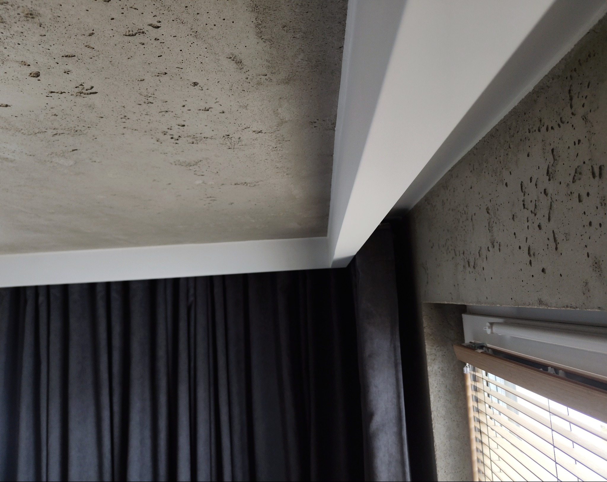 Szary sufit z fakturą betonu, białe listwy wykończeniowe, fragment okna z żaluzjami i zasłony w ciemnym kolorze. Minimalistyczny design wnętrza.