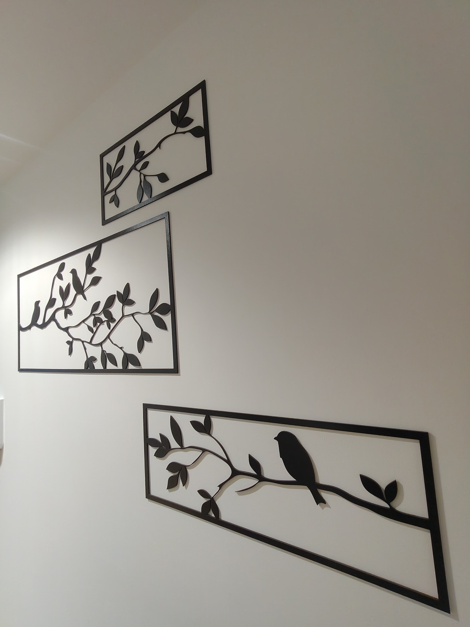 Dekoracja ścienna: trzy metalowe obrazy z motywem gałęzi i ptaków, rozmieszczone asymetrycznie na jasnej ścianie, tworzące nowoczesny wystrój wnętrza.