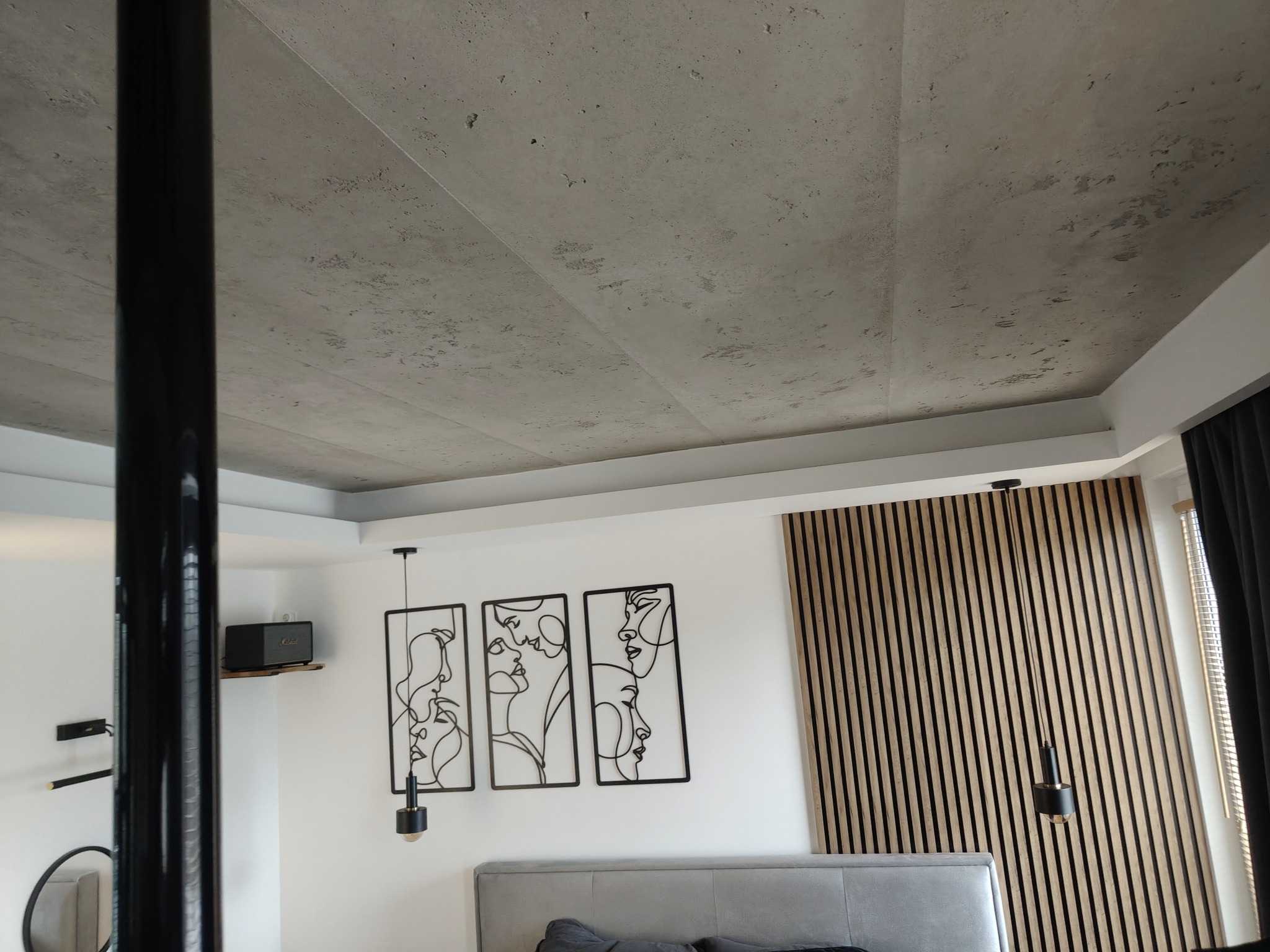 Betonowy sufit z minimalistycznymi lampami i dekoracjami ściennymi w nowoczesnym wnętrzu sypialni. Widoczna drewniana okładzina ścienna i elementy wyposażenia.