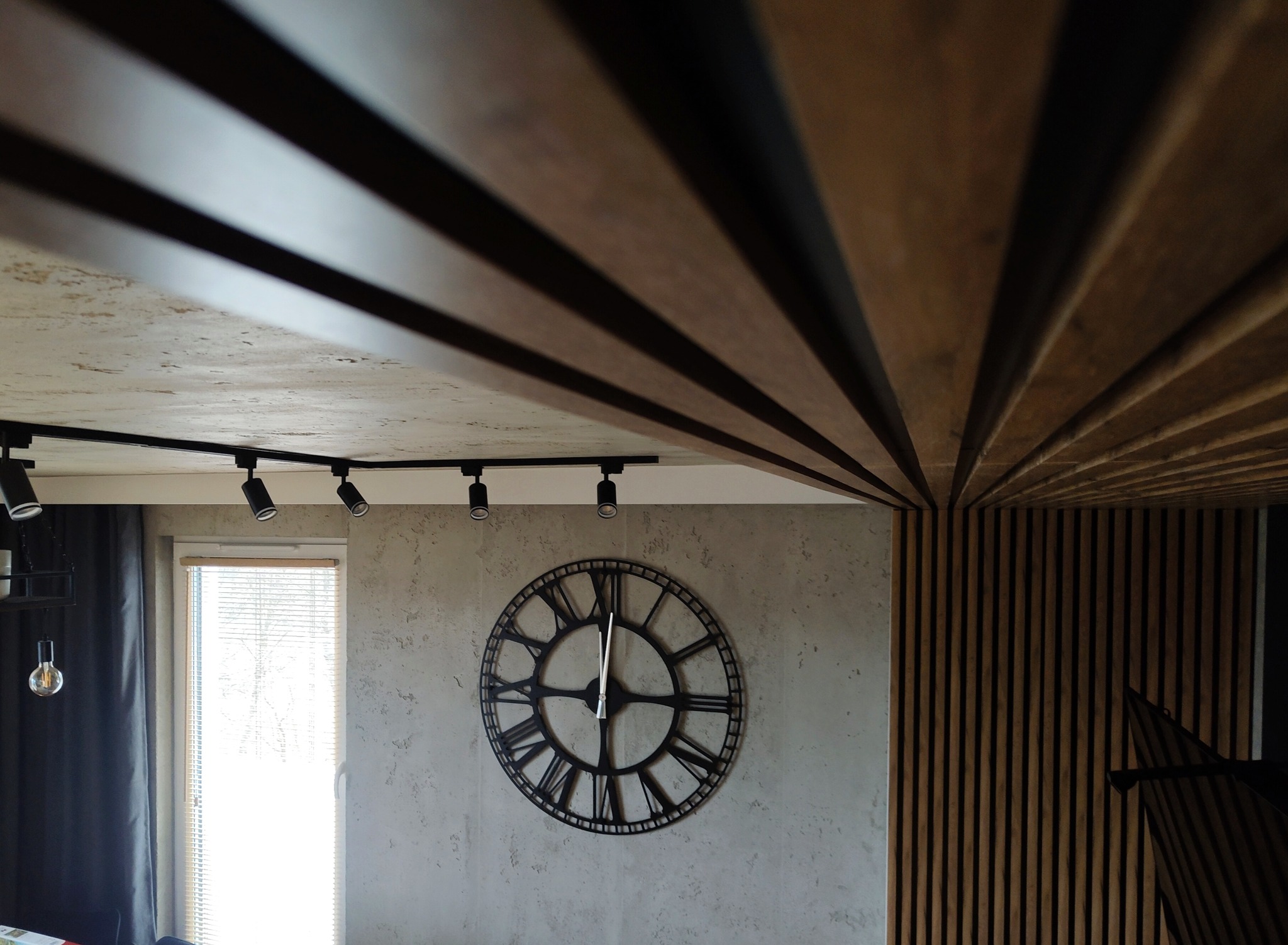 Nowoczesne wykończenie wnętrza: lamelki na suficie i ścianie, zegar na tle ściany o fakturze betonu, oświetlenie punktowe i okno z żaluzjami w tle.