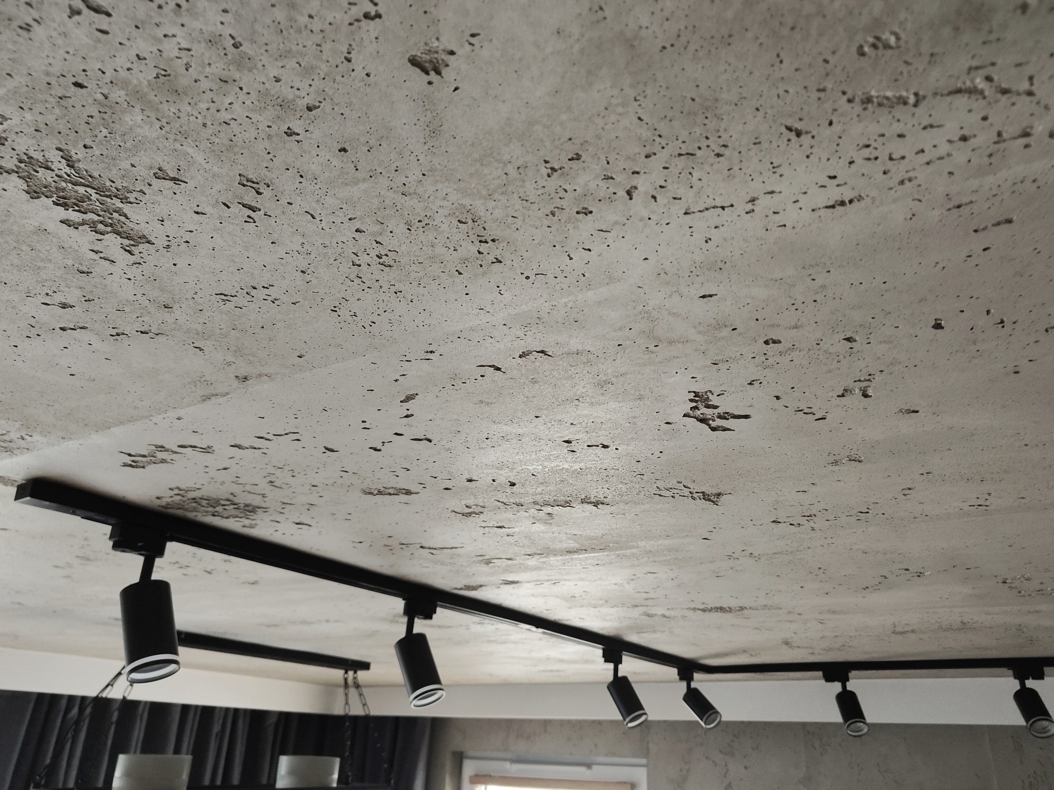 Stiukowy sufit z czarną szyną oświetleniową i reflektorami, widziany pod kątem. Wykończenie sufitu o nieregularnej fakturze, nowoczesne oświetlenie.