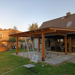 JS TIMBER JACEK SZCZYGIEŁ - Wznoszenie drewnianej pergoli na tarasie z kostki. Widoczne elementy konstrukcyjne, drabina i narzędzia. W tle dom z ceglaną elewacją i składowane drewno.