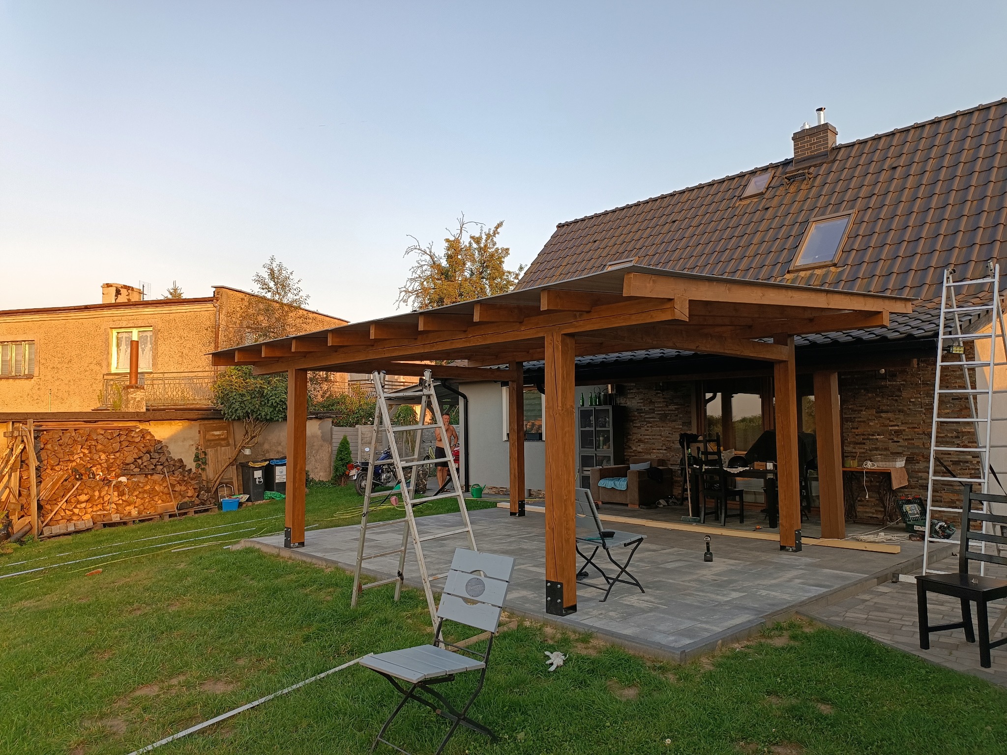 Wznoszenie drewnianej pergoli na tarasie z kostki. Widoczne elementy konstrukcyjne, drabina i narzędzia. W tle dom z ceglaną elewacją i składowane drewno.