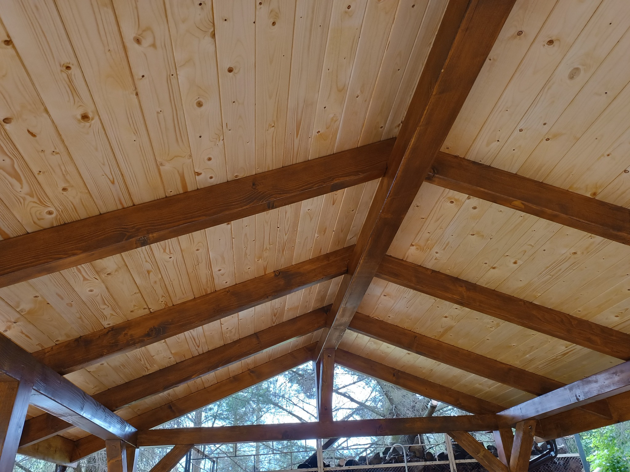 Drewniana konstrukcja altany z widocznym dachem z desek w jasnym odcieniu i ciemnobrązowymi belkami. Widok z dołu na konstrukcję i drzewa w tle.