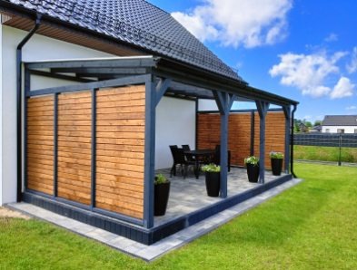 Drewniana pergola z ażurowymi ścianami, czarne donice z kwiatami, stół i krzesła w tle, kostka brukowa na podłożu, trawnik i błękitne niebo.