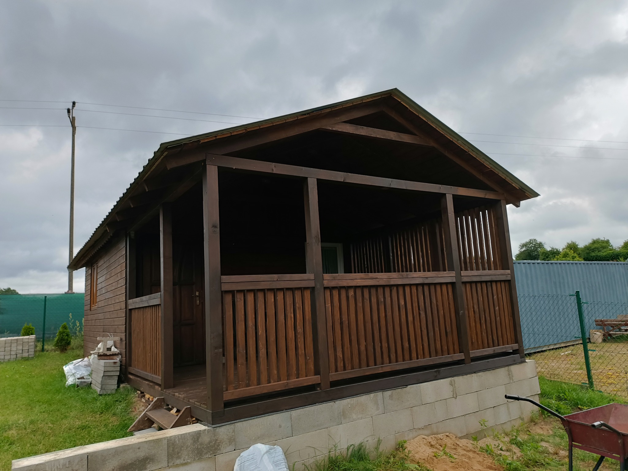 Ciemnobrązowa, drewniana altana z metalowym dachem, częściowo osłonięta, na fundamencie z bloczków betonowych, w tle pochmurne niebo i zieleń.
