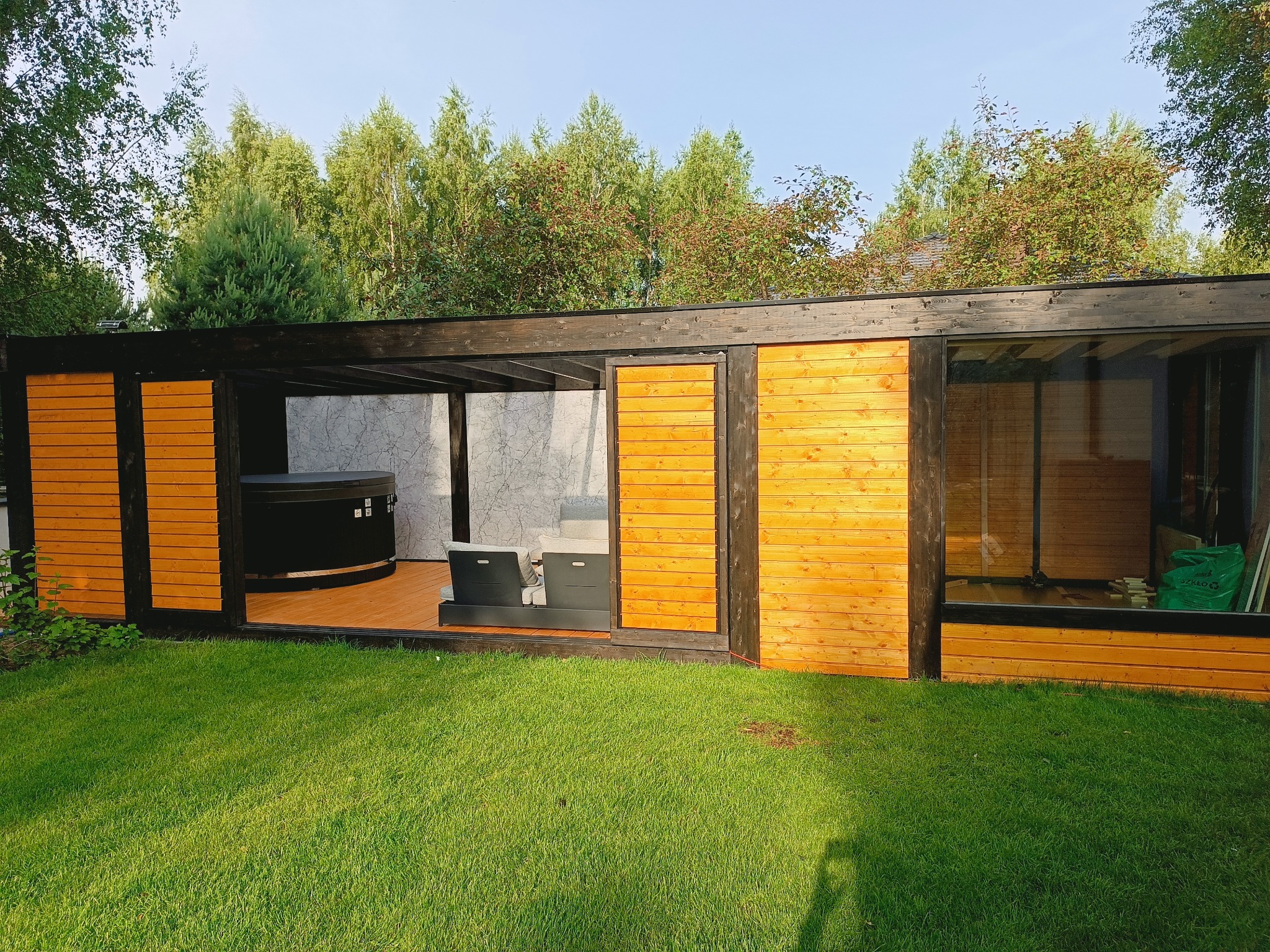 Nowoczesna altana z drewna i szkła z jacuzzi, otoczona zielonym trawnikiem. Widoczne elementy wykończeniowe i meble ogrodowe.