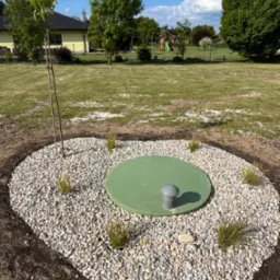 Okrągła rabata z białych kamieni, zielona pokrywa studzienki, małe krzewy i świeżo posadzone drzewko na tle zadbanego trawnika i placu zabaw.