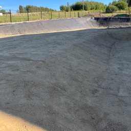 Zadbany Ogród - Przygotowany teren pod zbiornik wodny z wyłożoną folią, otoczony siatką ogrodzeniową i zielenią w tle, w słoneczny dzień. Widoczne ślady po pracy maszyn.