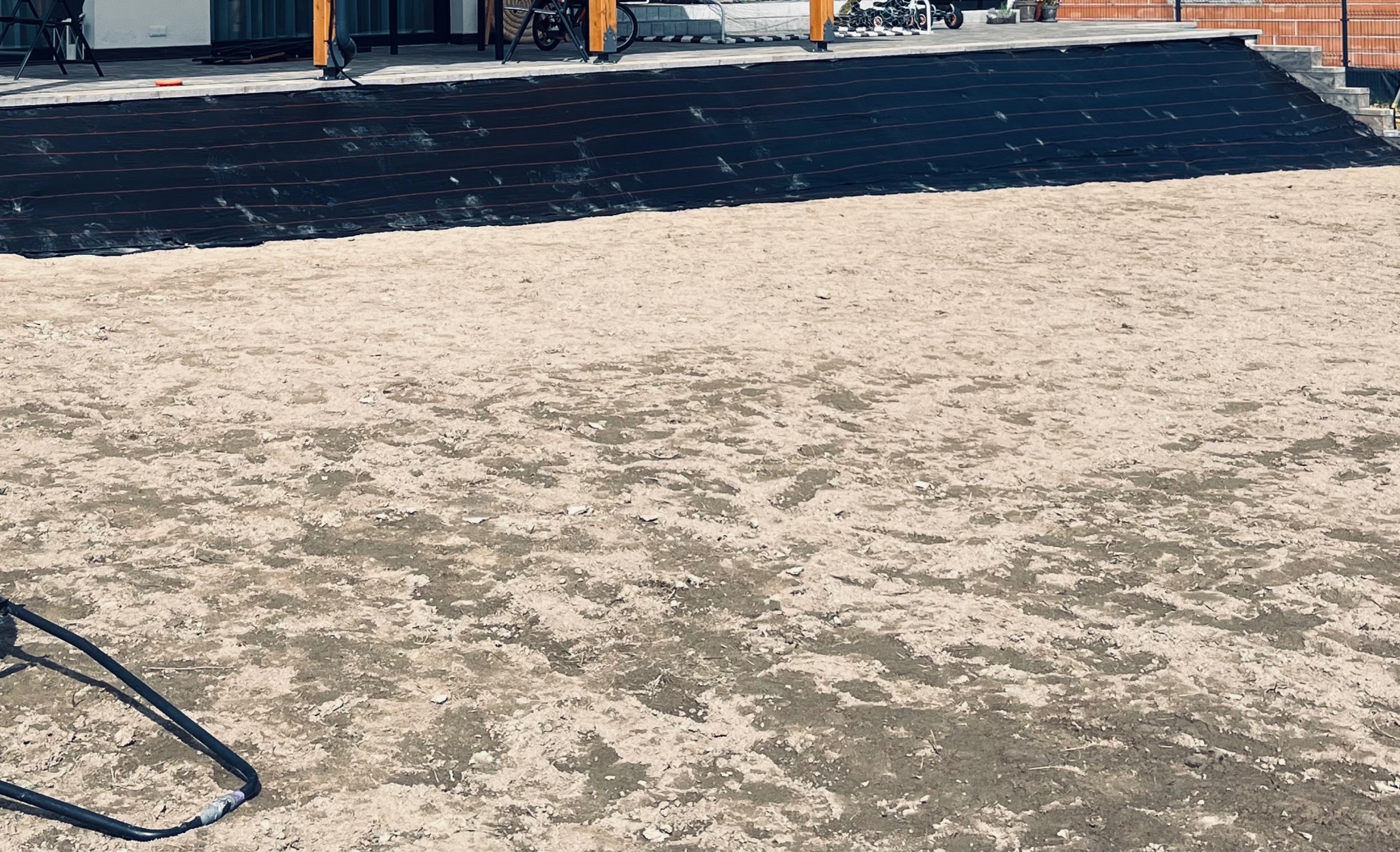 Przygotowanie terenu pod ogród: rozłożona czarna agrowłóknina na skarpie i wyrównana ziemia. Widoczny fragment tarasu i domu w tle, w słoneczny dzień.
