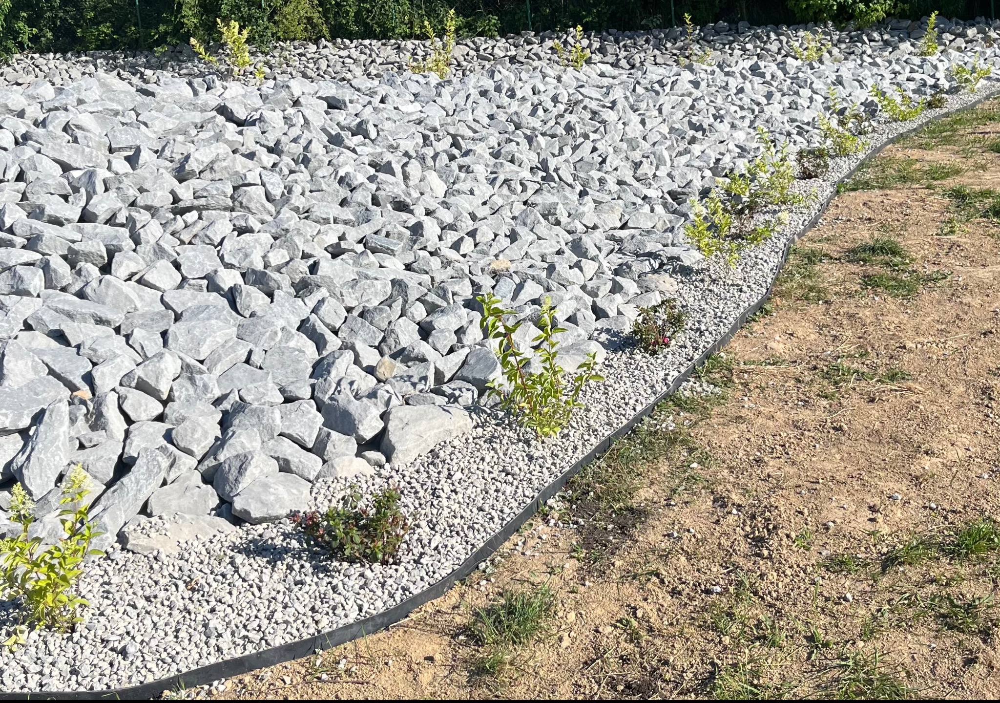 Kaskada z dużych, szarych kamieni i drobnego żwiru, oddzielona obrzeżem od ziemi. W tle zieleń drzew i krzewów. Nasadzenia roślin wzdłuż kamiennego brzegu.