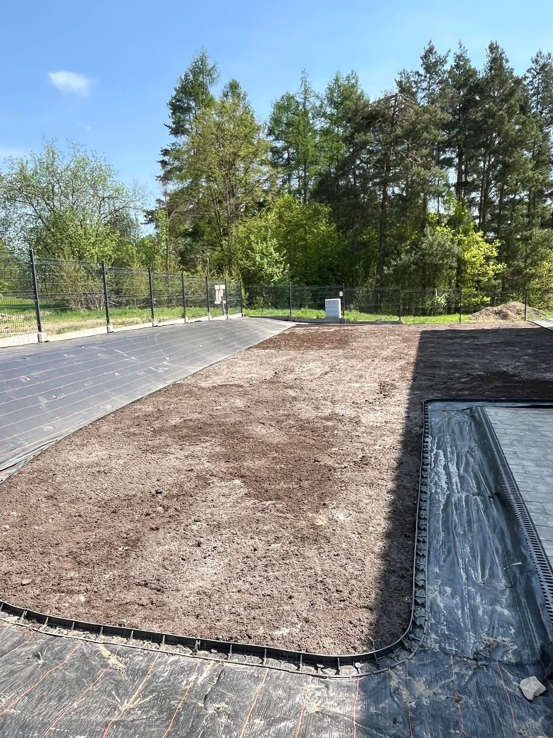 Przygotowany teren pod trawnik z rozłożoną agrowłókniną i obrzeżami trawnikowymi, widok z góry, słoneczny dzień. Część terenu z ziemią, część z agrowłókniną.