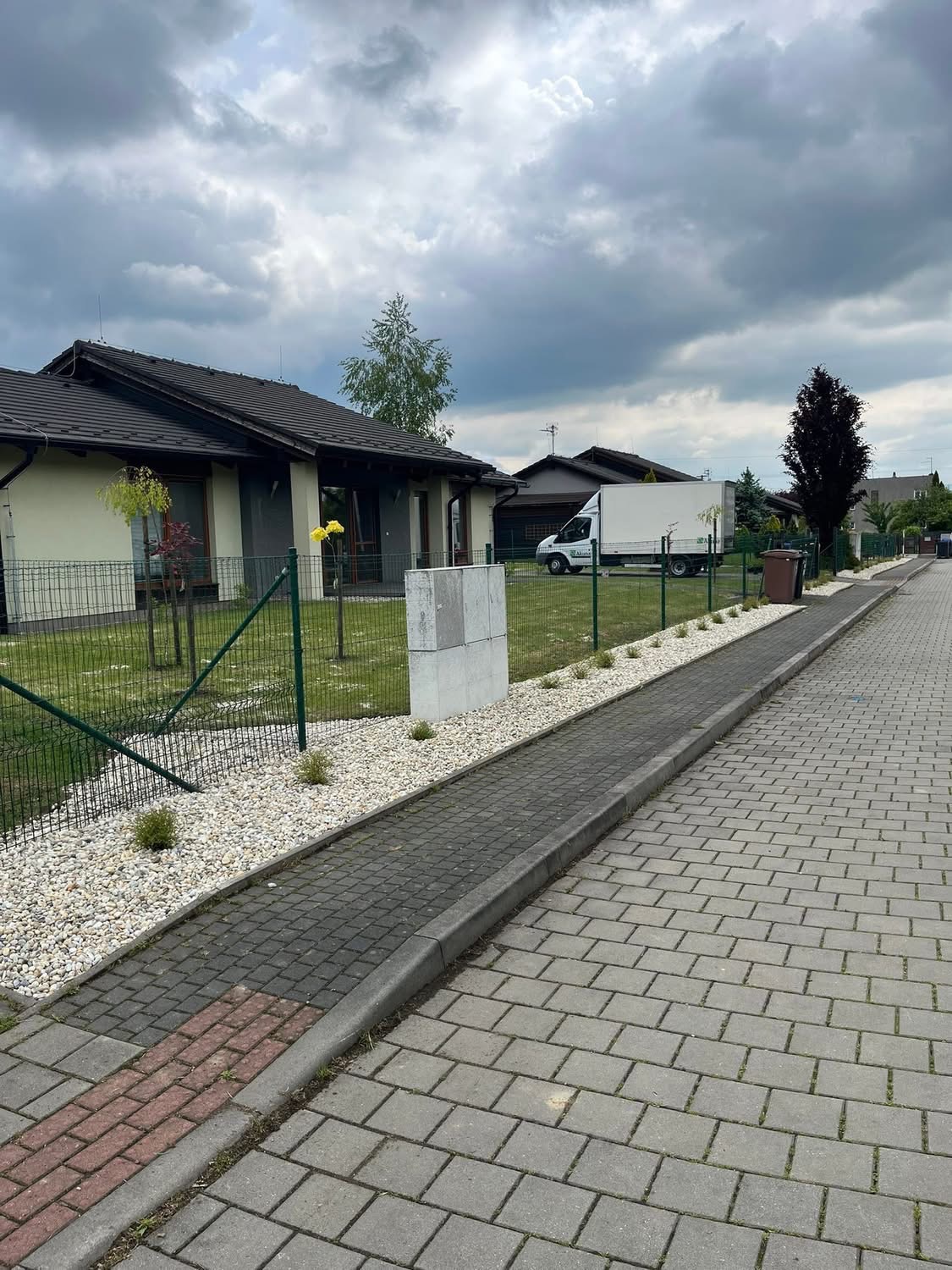 Ogród po realizacji: obsadzone kamieniami rabaty wzdłuż chodnika, trawnik za ogrodzeniem, dom jednorodzinny w tle i auto dostawcze. Pochmurne niebo.