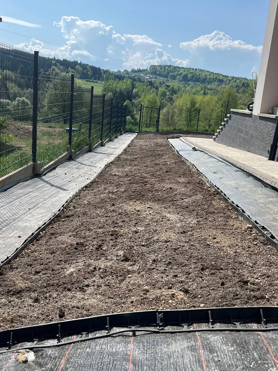 Przygotowany teren pod trawnik z widoczną czarną agrowłókniną i obrzeżami, w tle zieleń wzgórz i błękitne niebo. Etap prac przed założeniem trawnika.