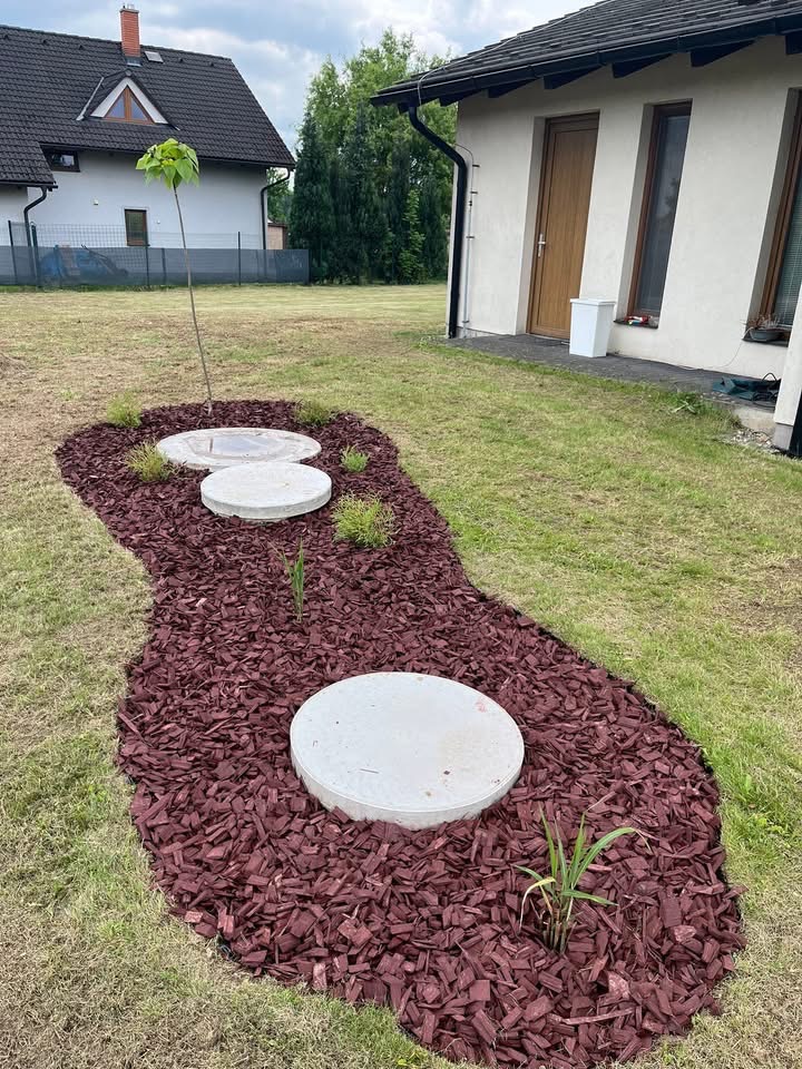 Nowo założony ogród z korą drzewną, okrągłymi pokrywami studzienek i młodym drzewkiem. Widok na dom z szarą elewacją i brązowym dachem.