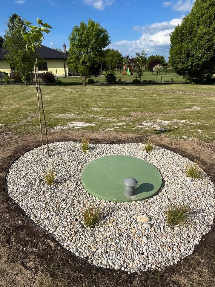 Okrągła studzienka kanalizacyjna w ogrodzie, otoczona białymi kamieniami i młodymi roślinami. W tle trawnik, drzewa i plac zabaw.