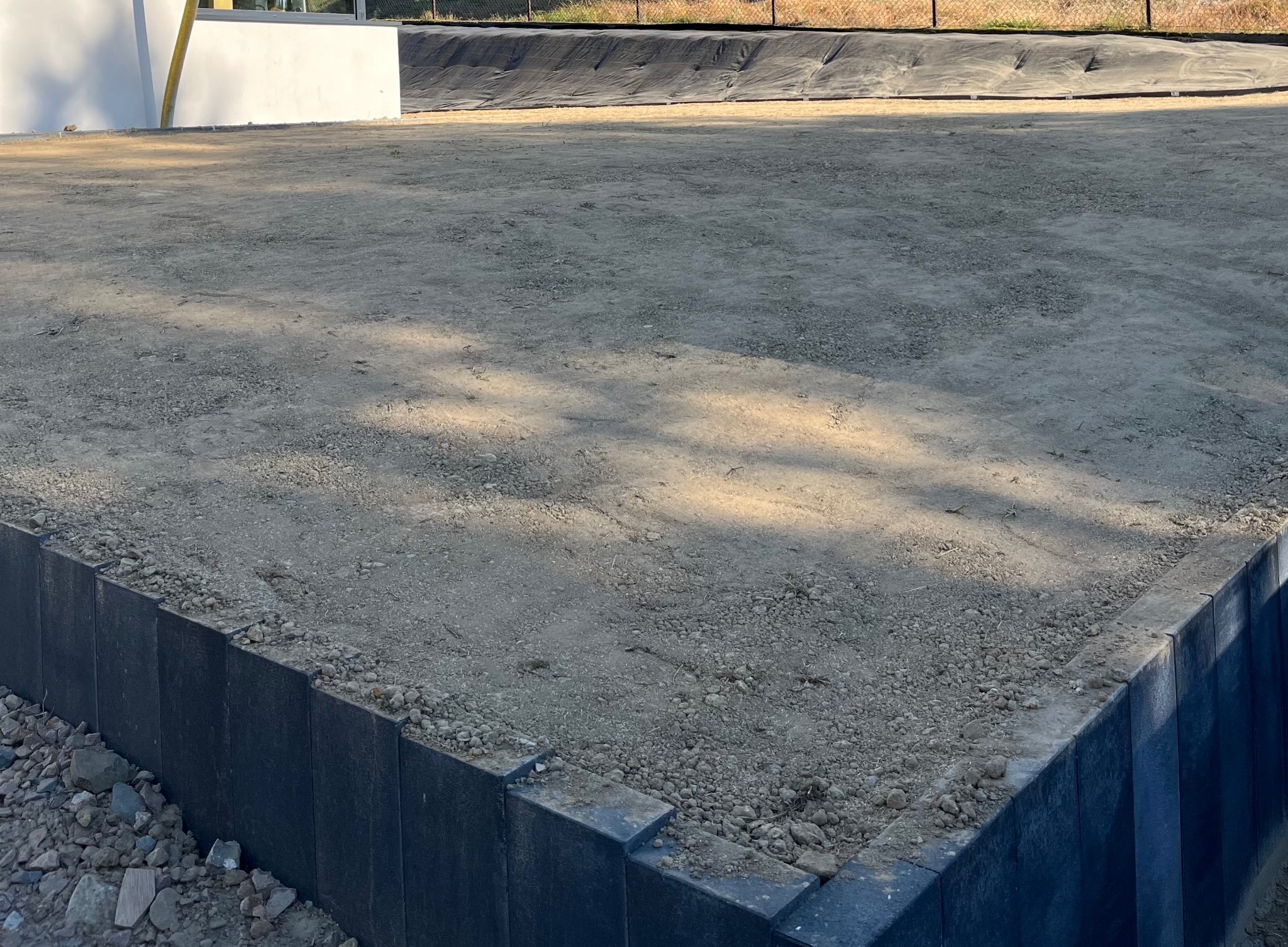 Przygotowany teren pod trawnik z obrzeżami z kostki. Widoczna ziemia, kamienie i czarne obrzeża. W tle folia zabezpieczająca. Gotowy teren pod trawnik.