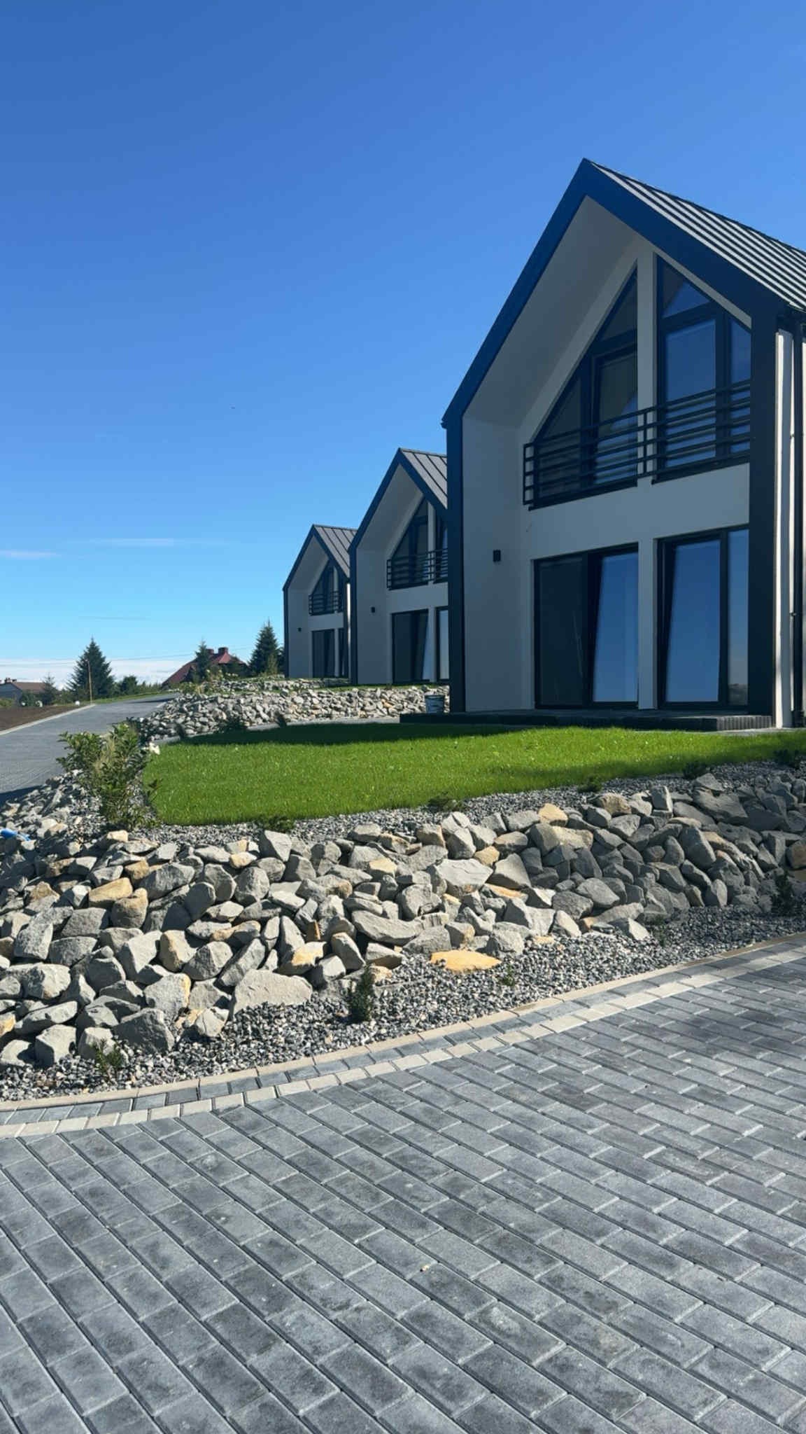 Nowoczesne domy z zadbanym trawnikiem i kamiennym nasypem. Kostka brukowa na pierwszym planie. Słoneczny dzień, błękitne niebo. Minimalistyczna architektura.