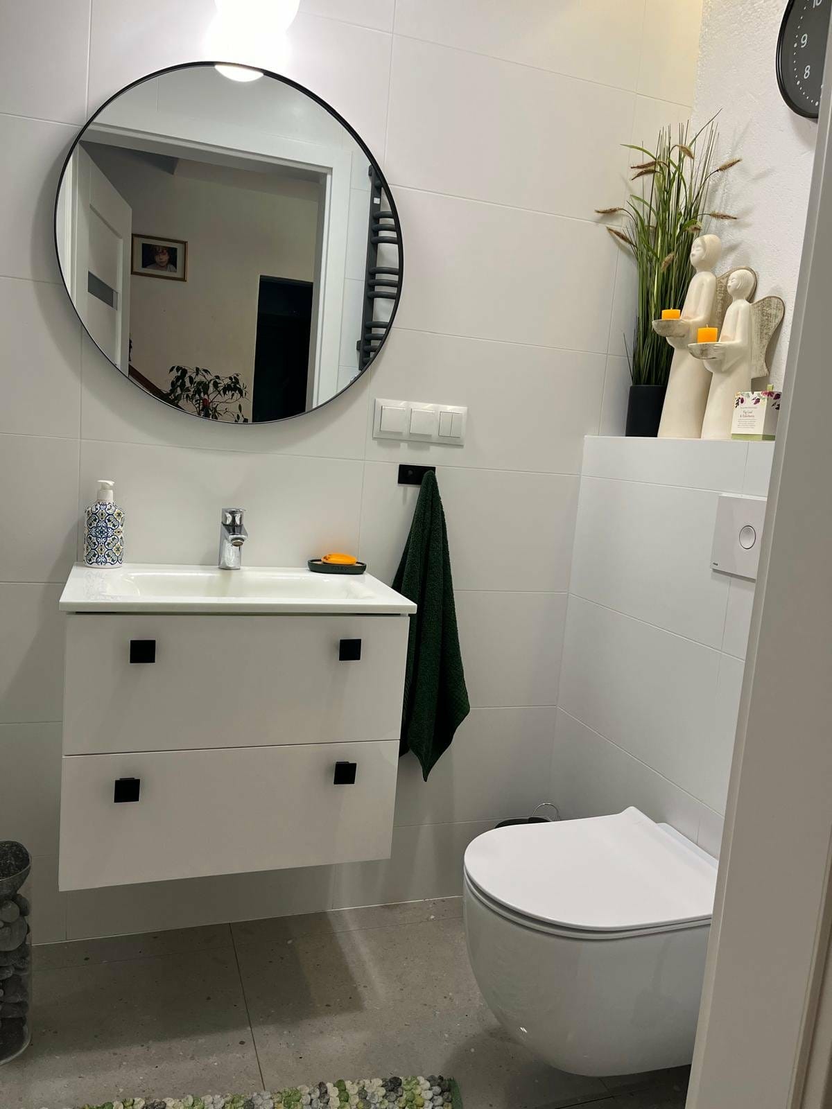 Nowoczesna łazienka z okrągłym lustrem, białą szafką pod umywalką z czarnymi uchwytami i wiszącą toaletą. Dekoracje: figurki aniołów i roślina w doniczce.
