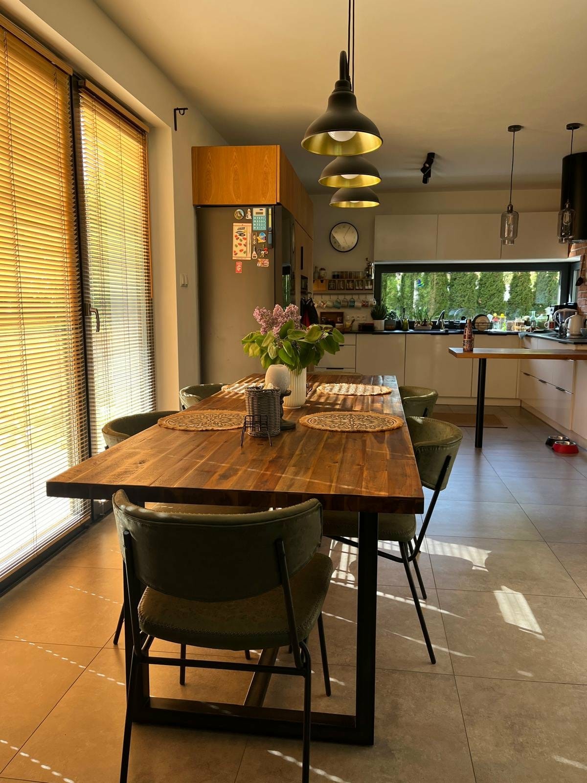 Drewniany stół z krzesłami w jasnej kuchni z dużym oknem i wiszącymi lampami. Na stole dekoracje i podkładki. Widok na ogród przez okno.