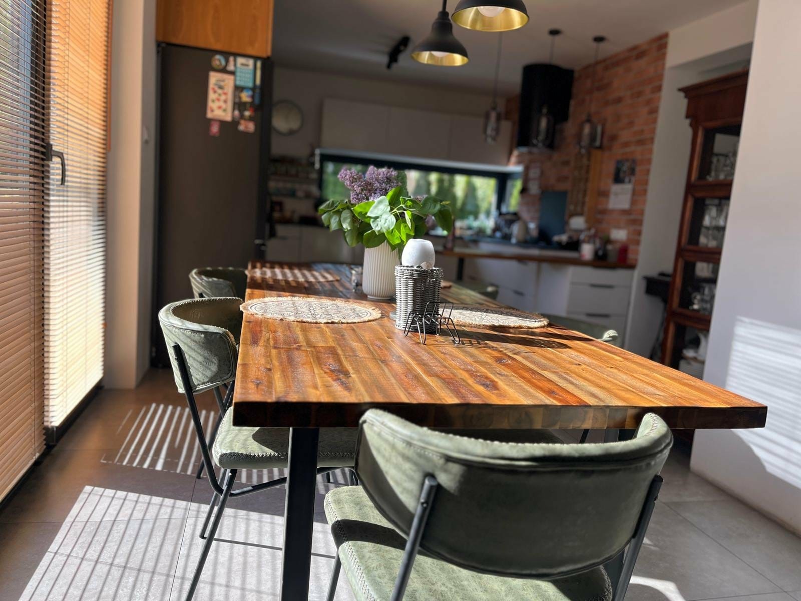 Rustykalny stół z drewnianym blatem i zielonymi krzesłami w słonecznej jadalni. W tle kuchnia z ceglaną ścianą i dekoracje kwiatowe na stole.