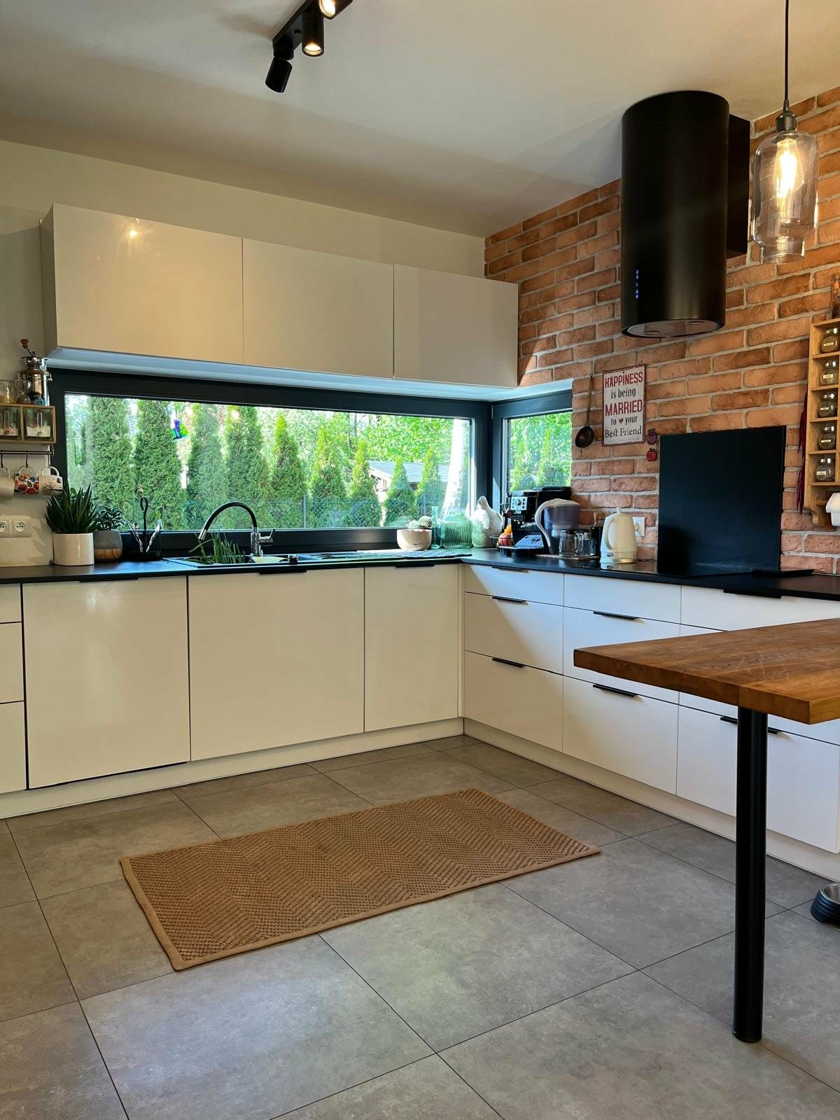 Nowoczesna kuchnia z białymi szafkami, czarnym blatem i ceglaną ścianą. Duże okno z widokiem na zieleń. Na podłodze szary gres i dywanik.