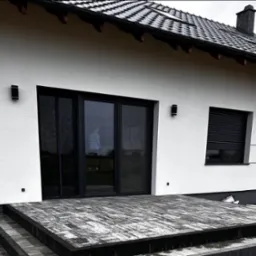 Nowoczesny taras z kostki brukowej w odcieniach szarości, prowadzący do domu z białymi ścianami i czarnymi oknami. Widoczne schody z kostki i fragment ogrodu.