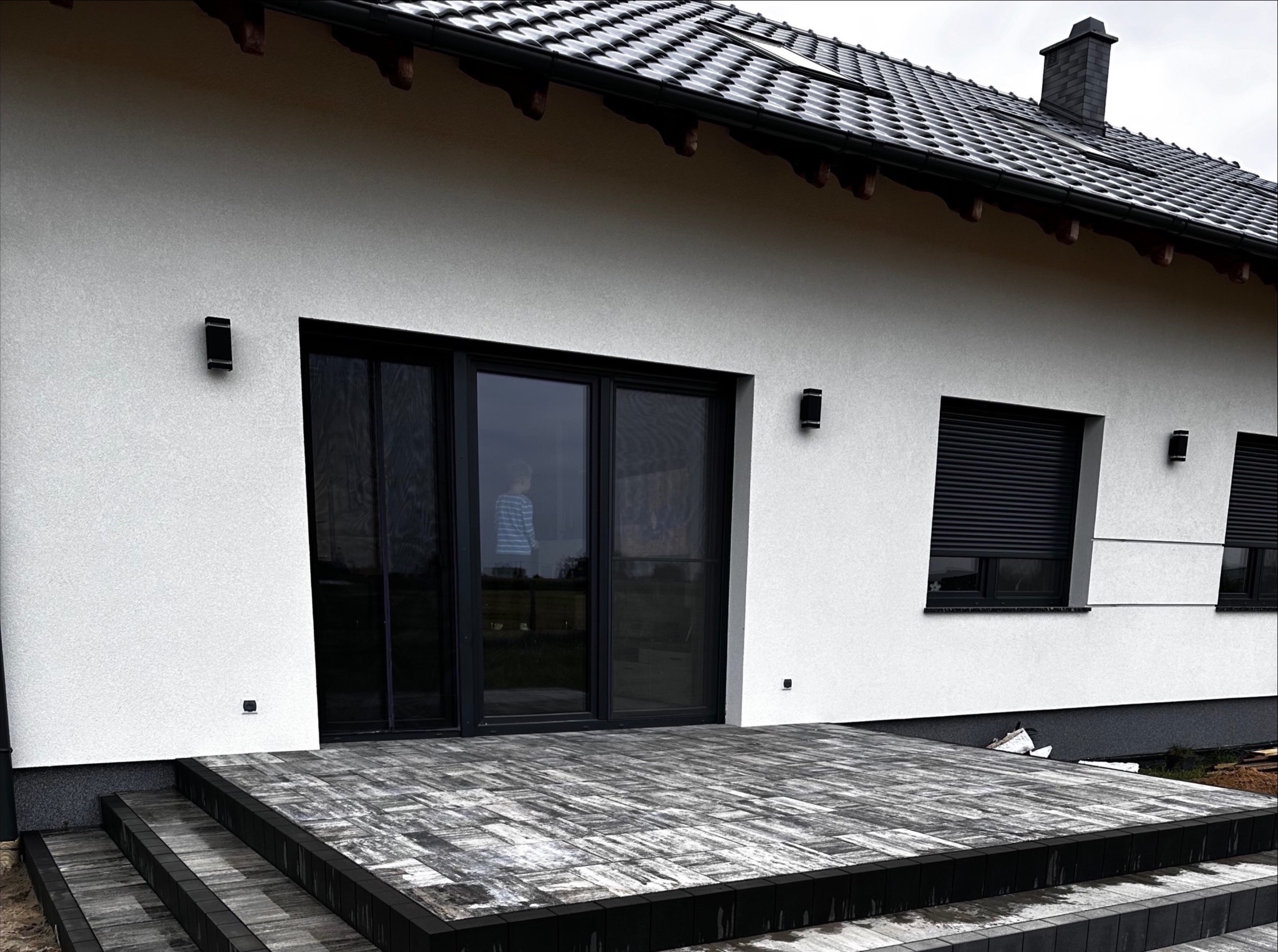 Nowoczesny taras z kostki brukowej w odcieniach szarości, prowadzący do domu z białymi ścianami i czarnymi oknami. Widoczne schody z kostki i fragment ogrodu.