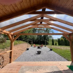 Zadaszona, drewniana altana z transparentnym dachem. Widok na ogród z miejscem na ognisko i relaks. Na pierwszym planie drewniany podest i dekoracyjny dywan.
