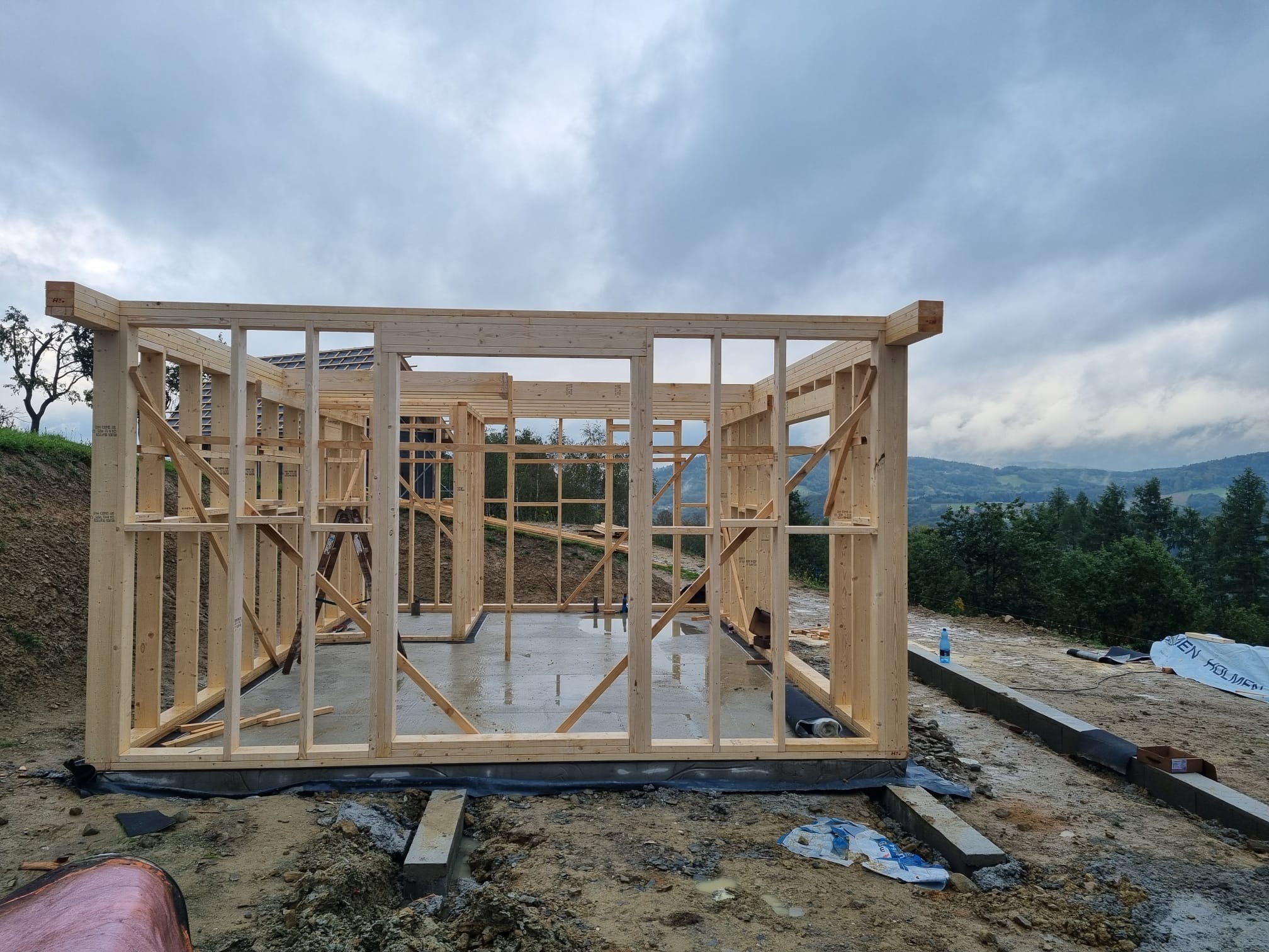 Surowa konstrukcja domu szkieletowego z widocznym betonowym fundamentem na tle pochmurnego nieba i zalesionych wzgórz. Widoczne detale łączeń drewnianych elementów.