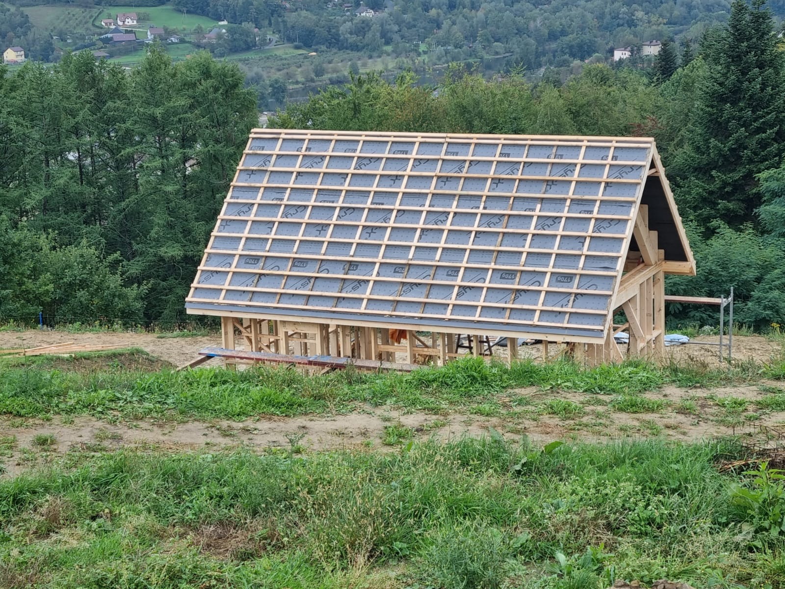 Konstrukcja szkieletowa domu w trakcie budowy, widok z zewnątrz na drewniany szkielet i wstępnie pokryty dach na tle zieleni i wzgórz.