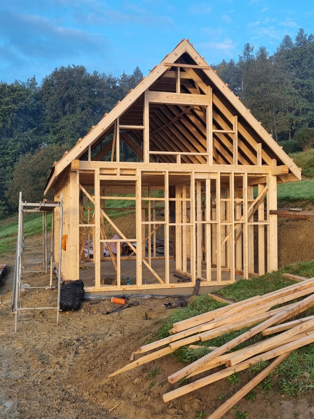 Wznoszenie szkieletu drewnianego domu na fundamencie, widoczne elementy konstrukcyjne ścian i dachu, z belkami na ziemi. W tle zieleń drzew i błękitne niebo.