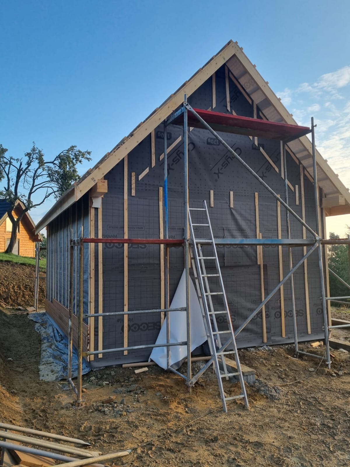 Budowa domu szkieletowego w trakcie: widoczne rusztowanie, drewniane elementy konstrukcyjne i czarna membrana. W tle fragment drugiego domu.