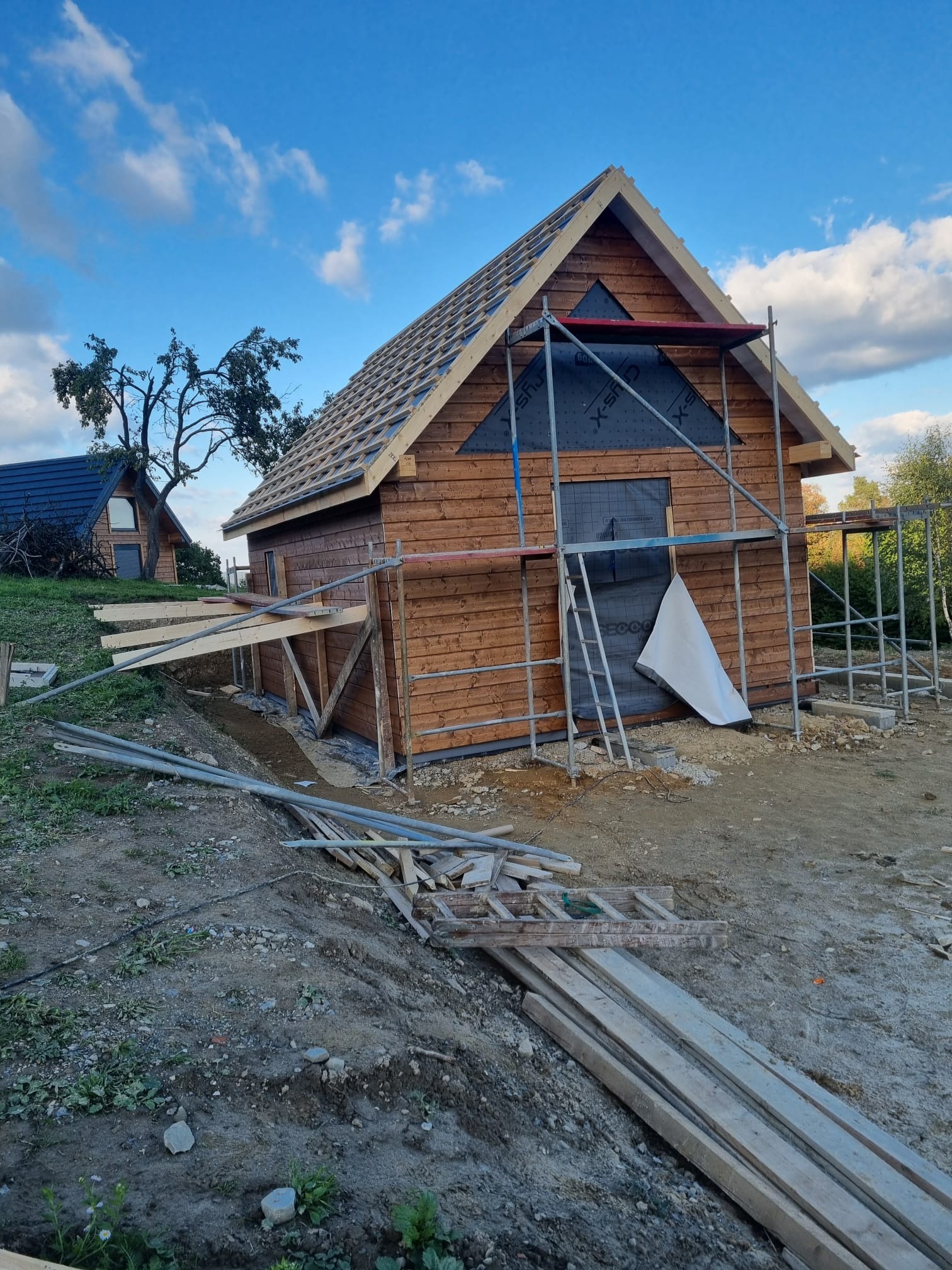 Brązowy, drewniany dom w trakcie budowy, z widocznym rusztowaniem, membraną na ścianie i elementami konstrukcyjnymi dachu. W tle drugi budynek i błękitne niebo.
